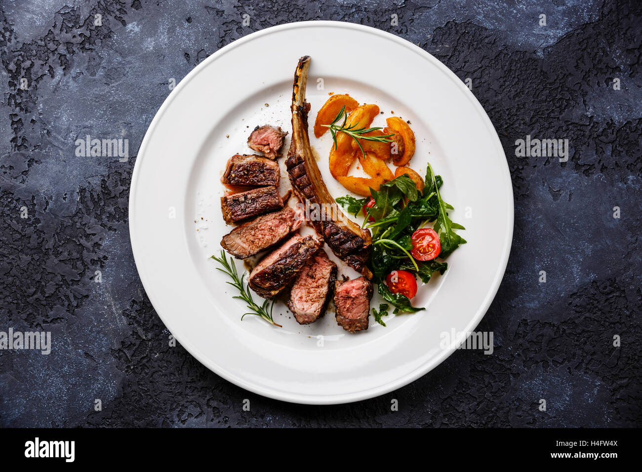 Fette di manzo alla griglia la nervatura sul tessuto osseo su piastra bianca con spicchi di patate e insalata con pomodori e rucola su sfondo scuro Foto Stock