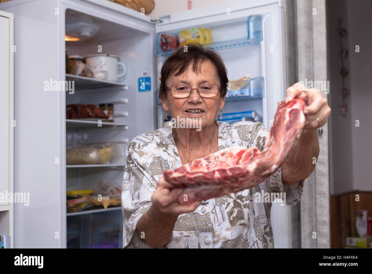 Senior donna azienda materie costolette di maiale mentre in piedi di fronte ad aprire il frigorifero in cucina. Foto Stock