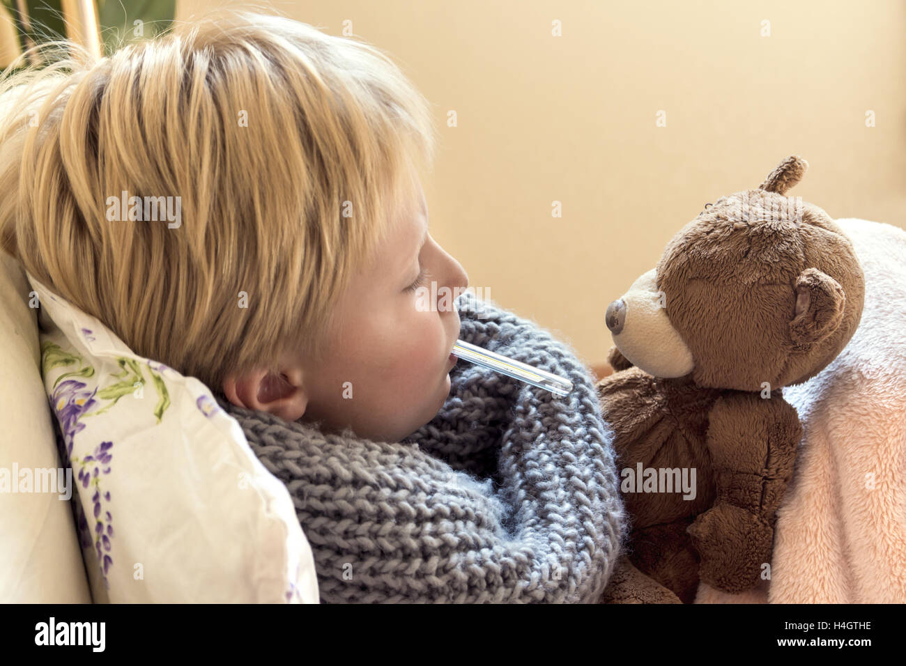 Bambino malato (ragazzo biondo, capretto) giacente a letto con il termometro e orsacchiotto Foto Stock