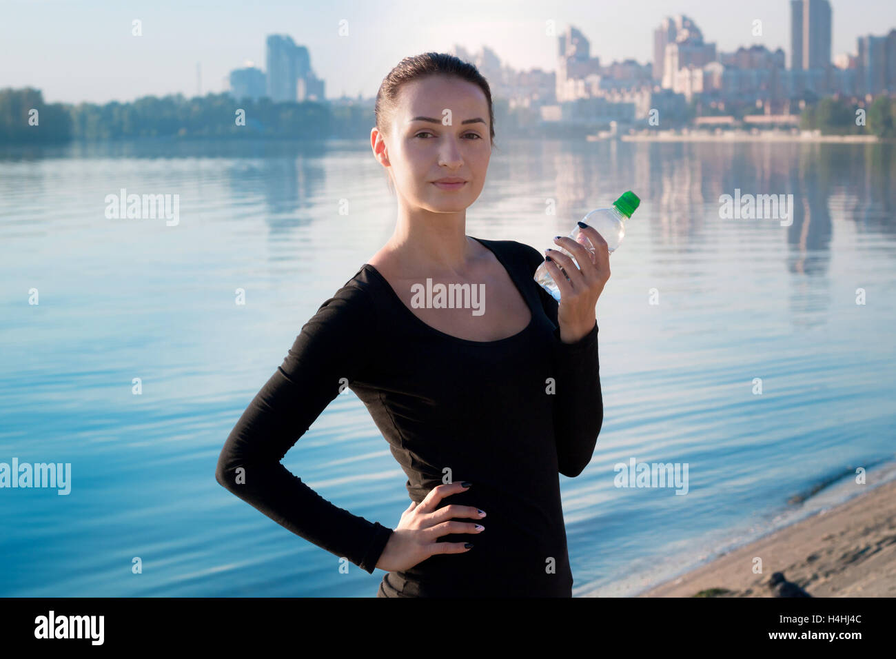 Giovane bella donna fitness detiene una bottiglia d'acqua al mattino il fiume e la città sul backround Foto Stock