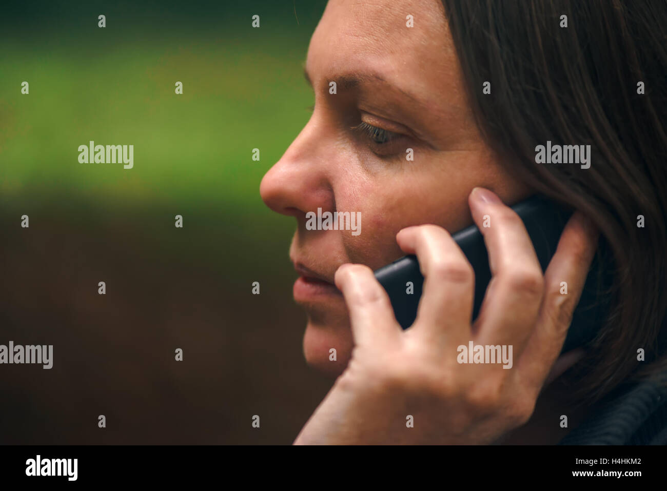Donna con espressione seria parlando al telefono mobile nel parco, drammatica luce proveniente attraverso le cime degli alberi e cadere sul suo viso Foto Stock Donna con espressione seria parlando al telefono mobile nel parco, drammatica luce proveniente attraverso le cime degli alberi e cadere sul suo viso Foto Stock