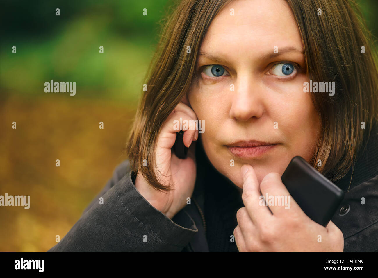 Donna con espressione seria parlando al telefono mobile nel parco, drammatica luce proveniente attraverso le cime degli alberi e cadere sul suo viso Foto Stock Donna con espressione seria parlando al telefono mobile nel parco, drammatica luce proveniente attraverso le cime degli alberi e cadere sul suo viso Foto Stock