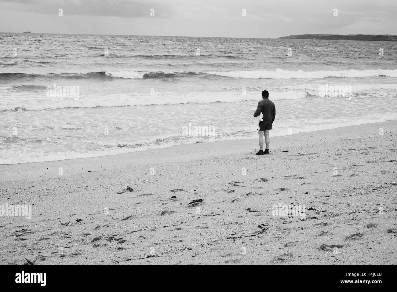 Uomo in un cappello sorge accanto al mare con un asciugamano sotto il suo braccio come onde venire in a Falmouths Gyllyngvase Beach 15-10-16 Foto Stock