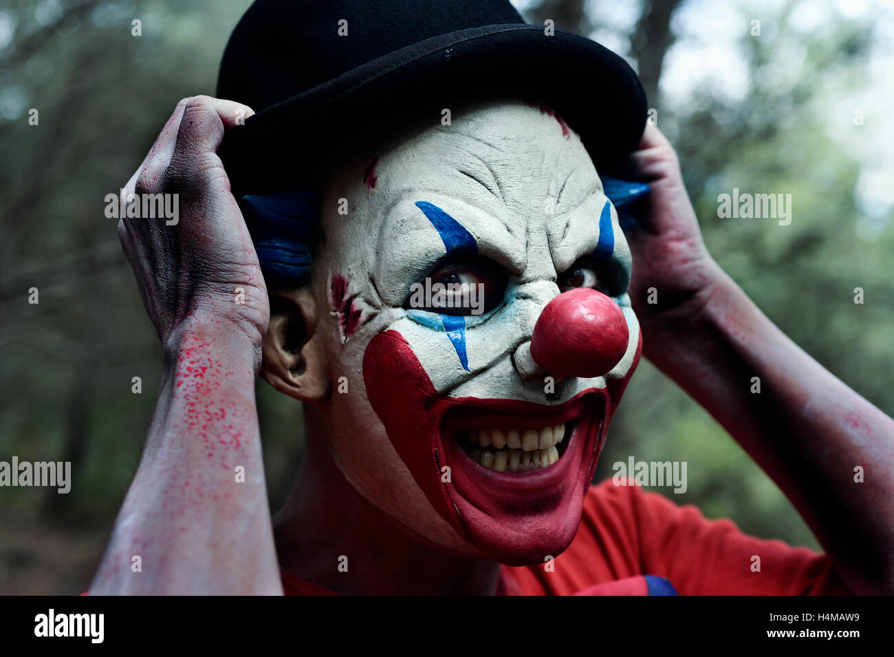 Primo piano di una paura male clown nei boschi sorridere mentre si adegua il suo cappello bowler Foto Stock