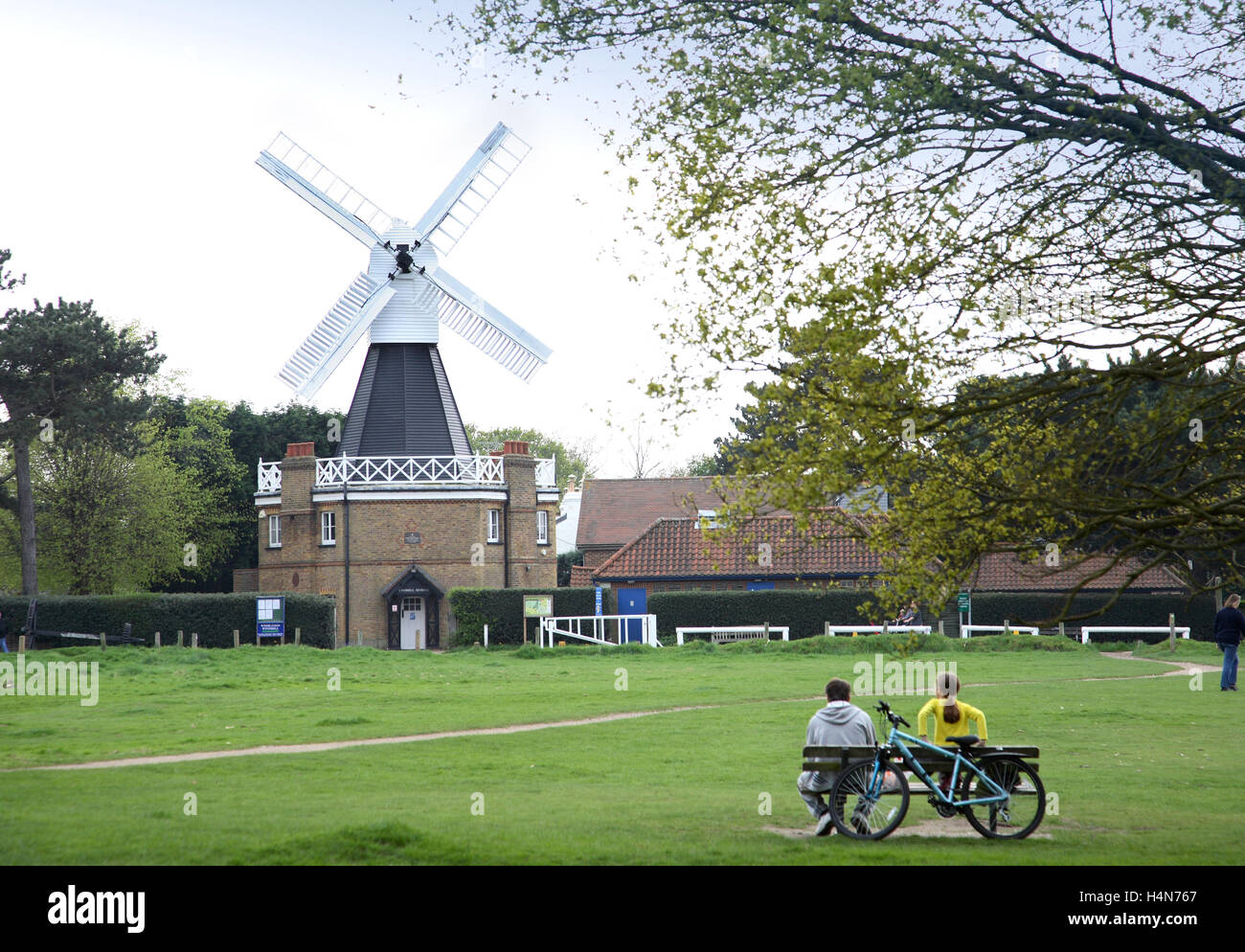 Il mulino a vento su Wimbledon Common a South London, Regno Unito, ora un museo locale di storia locale. La coppia SA si siede su una panchina con la bicicletta appoggiata dietro. Foto Stock