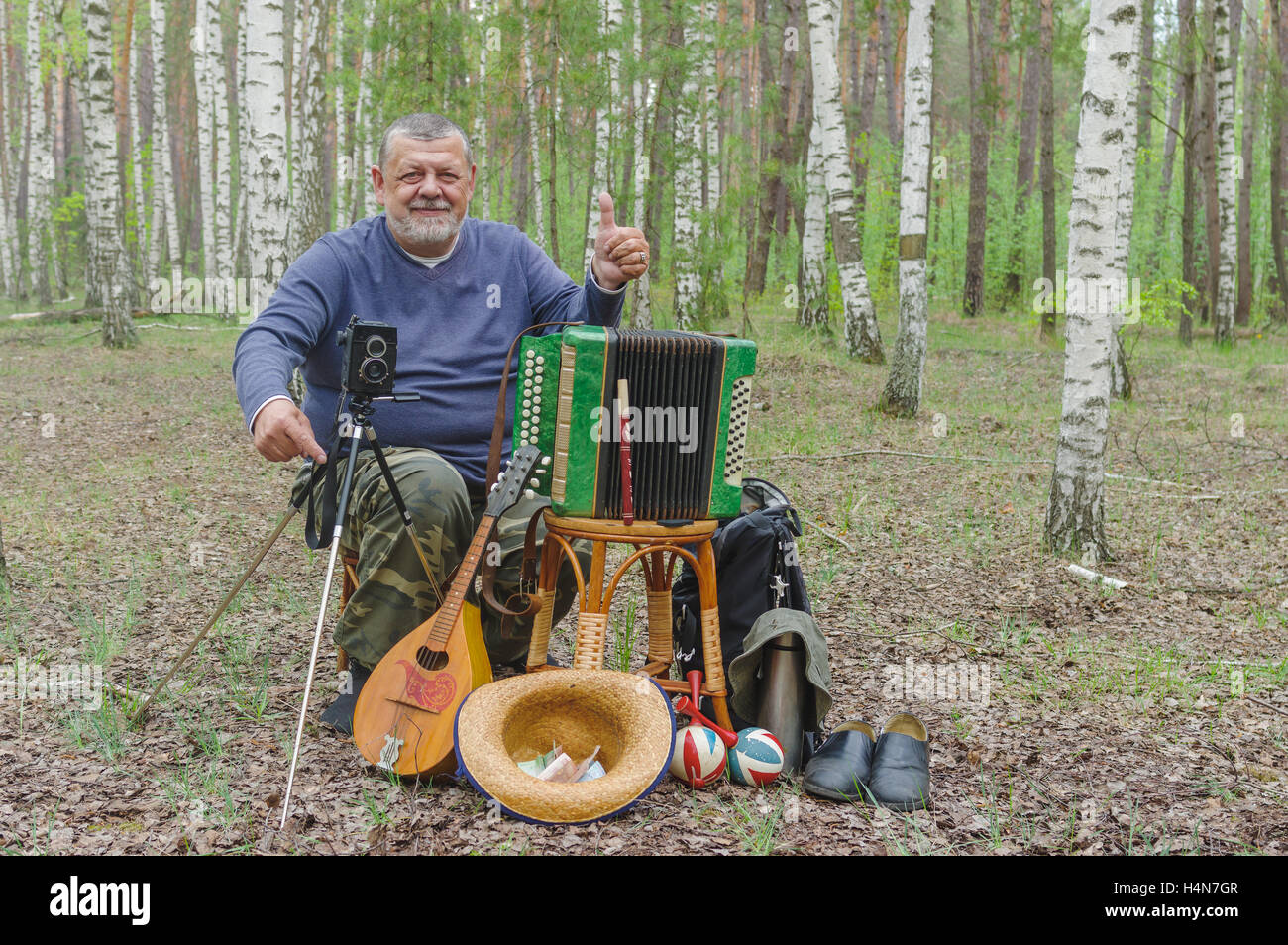 Felice senior camper è di riposo nella foresta di betulla, seduti su uno sgabello in vimini e tenendo il mandolino Foto Stock