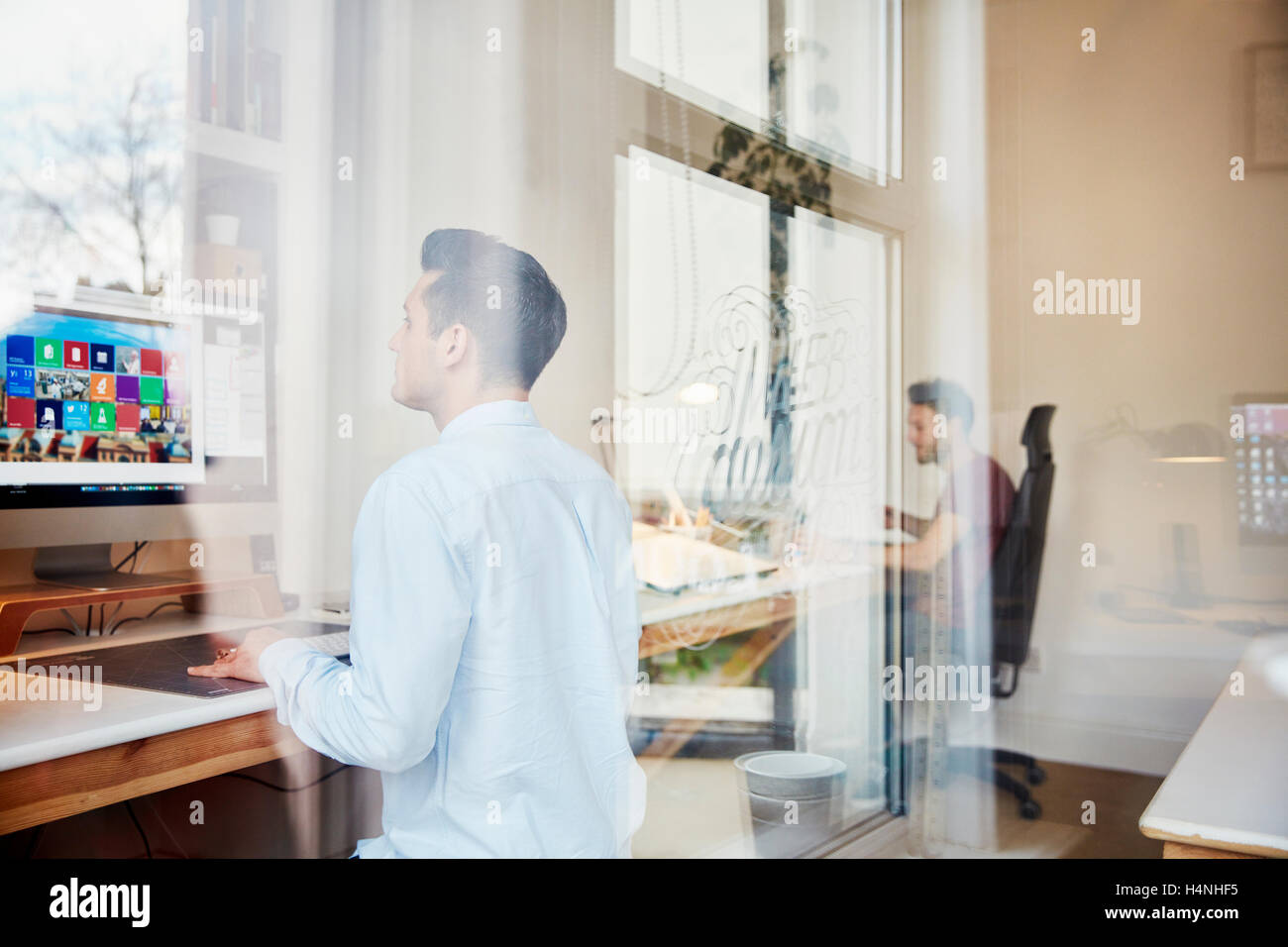 Due uomini al lavoro sul computer in uno studio di design, vista attraverso una finestra e riflessioni del telaio della finestra e alberi. Foto Stock