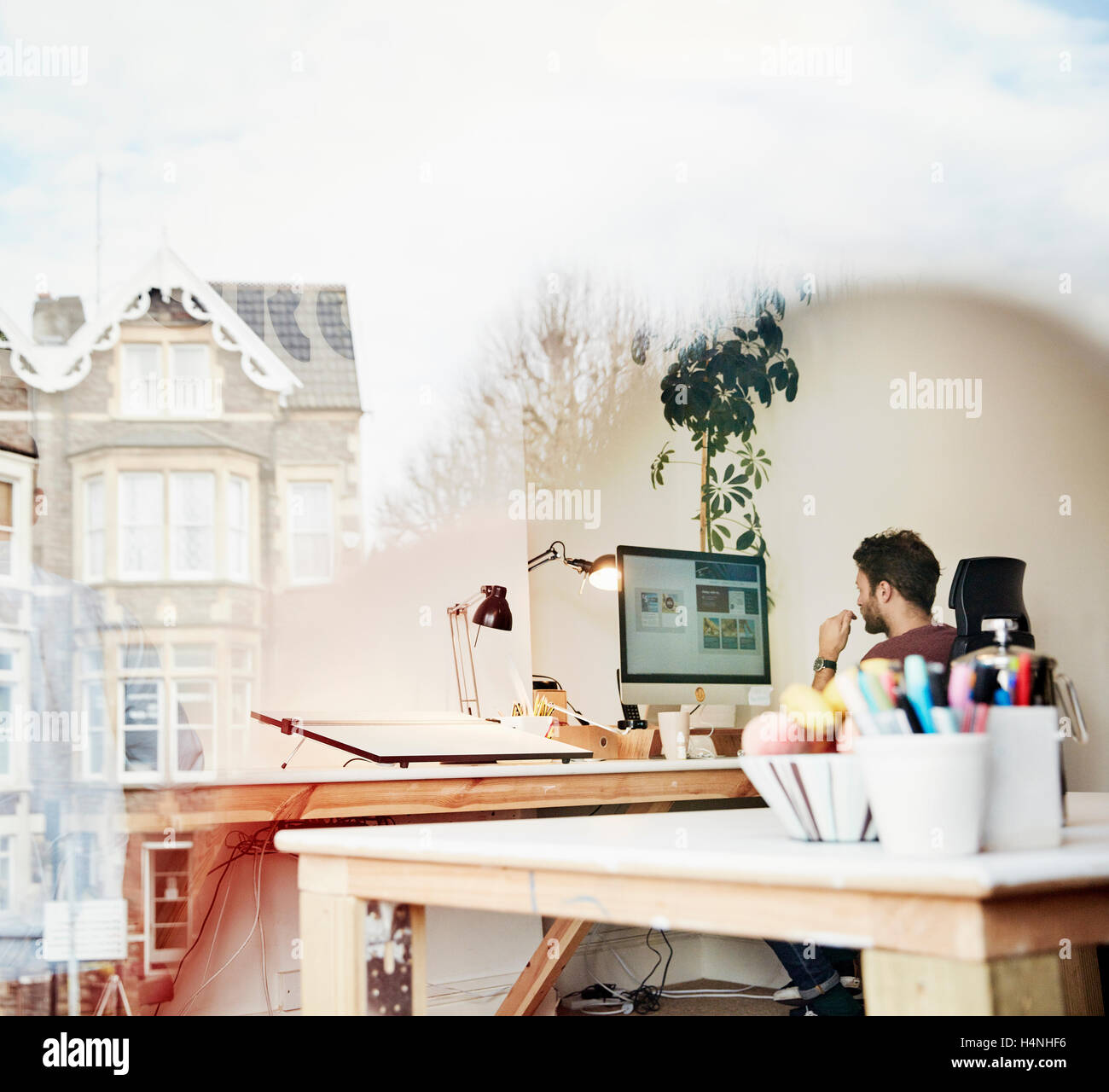 Un uomo che lavora a uno schermo di computer in uno studio di design. Vista attraverso la finestra, riflessioni di edifici. Foto Stock