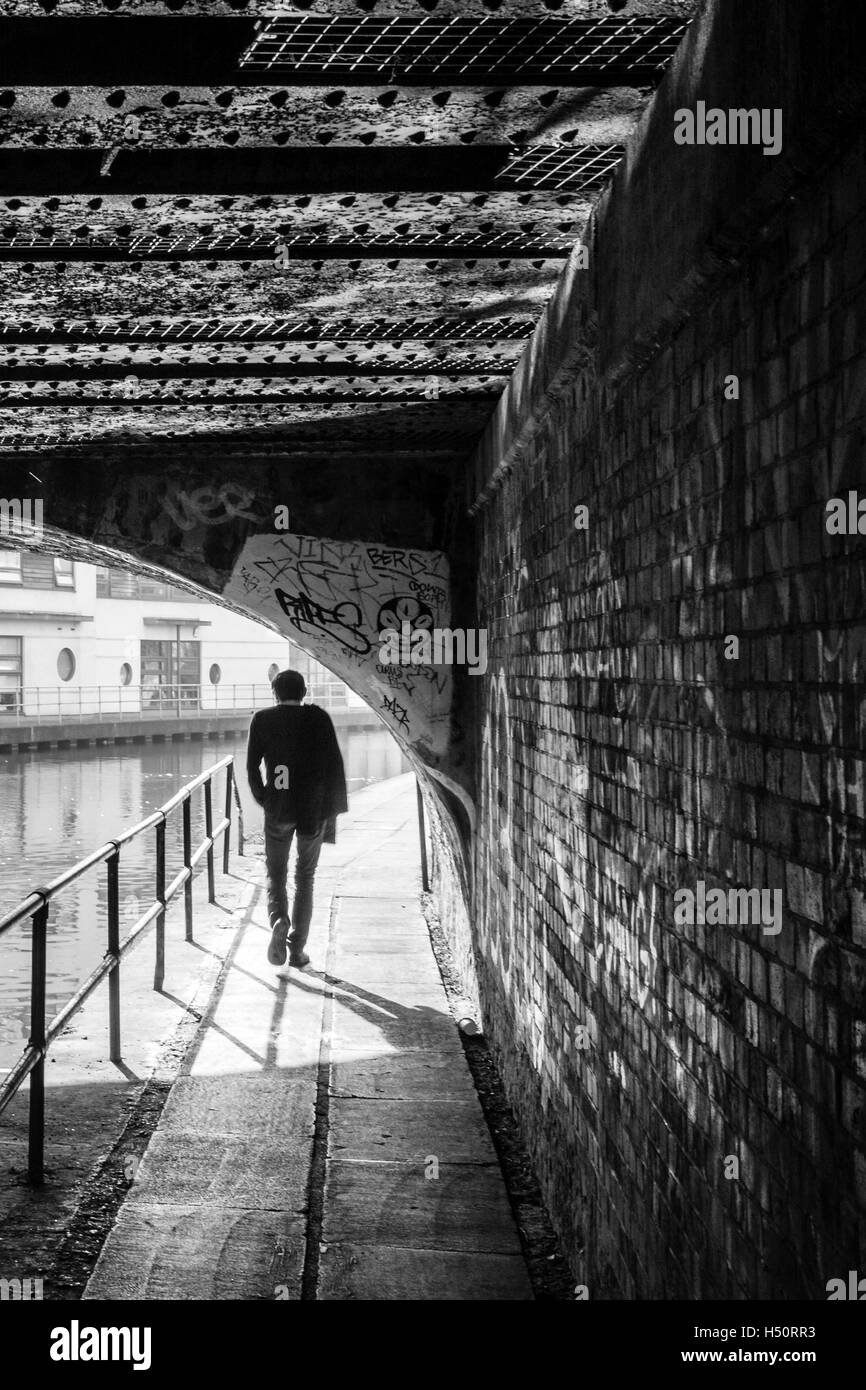 Immagine in bianco e nero di una singola persona camminando sotto un ponte sulla strada alzaia del Regent's Canal, stagliano e retroilluminato, London, Regno Unito Foto Stock