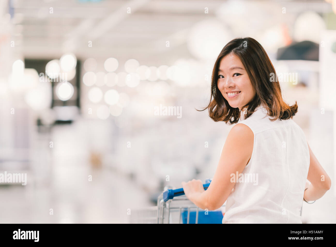 Bella giovane donna asiatica sorridente, con carrello, shopping center o department store in scena, sfocatura dello sfondo bokeh di fondo Foto Stock