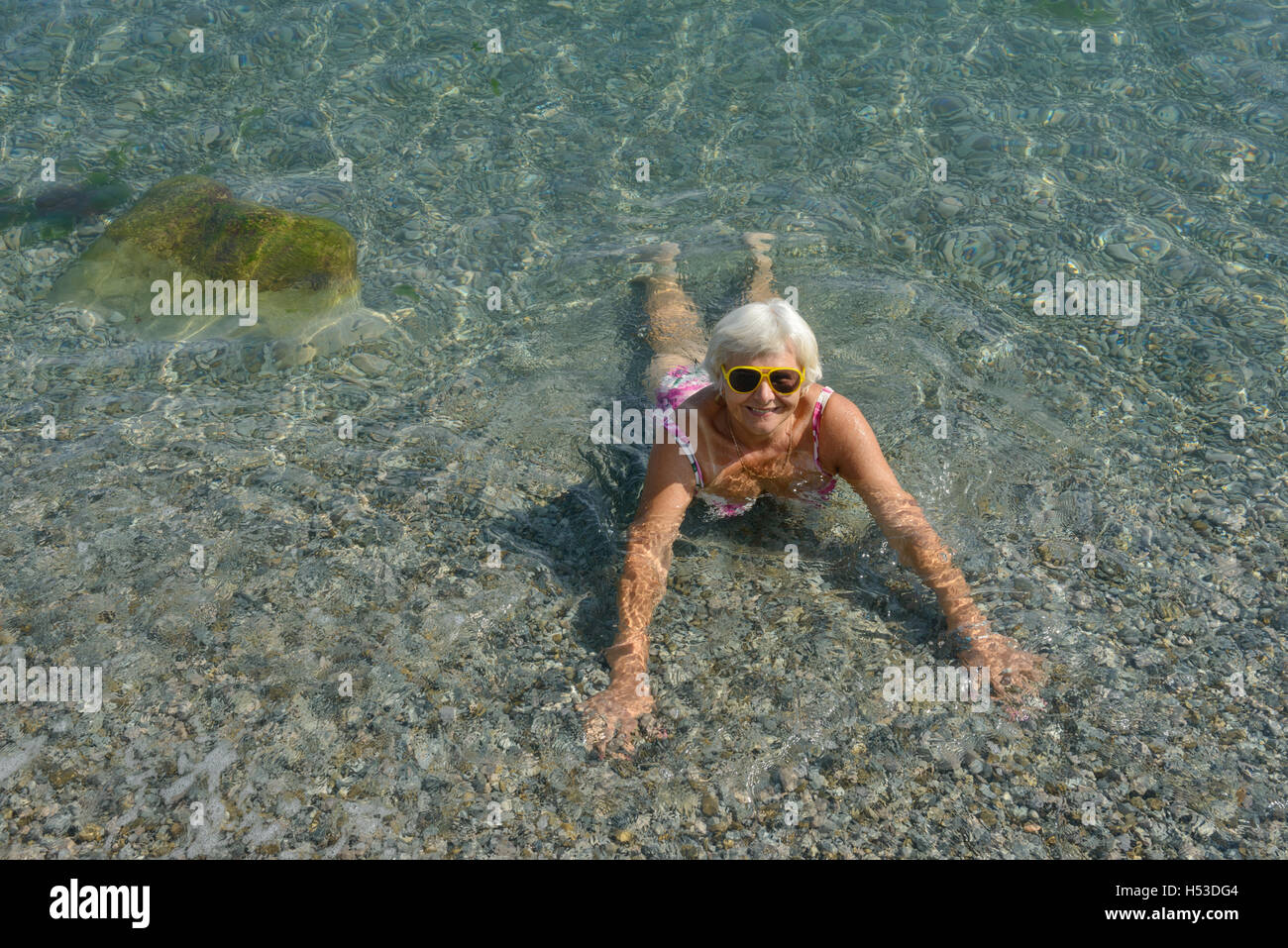 A piena lunghezza Ritratto di dama senior in occhiali da sole che la pronazione è chiaramente in acque trasparenti del mare ghiaiosa spiaggia vicino alle alghe cove Foto Stock