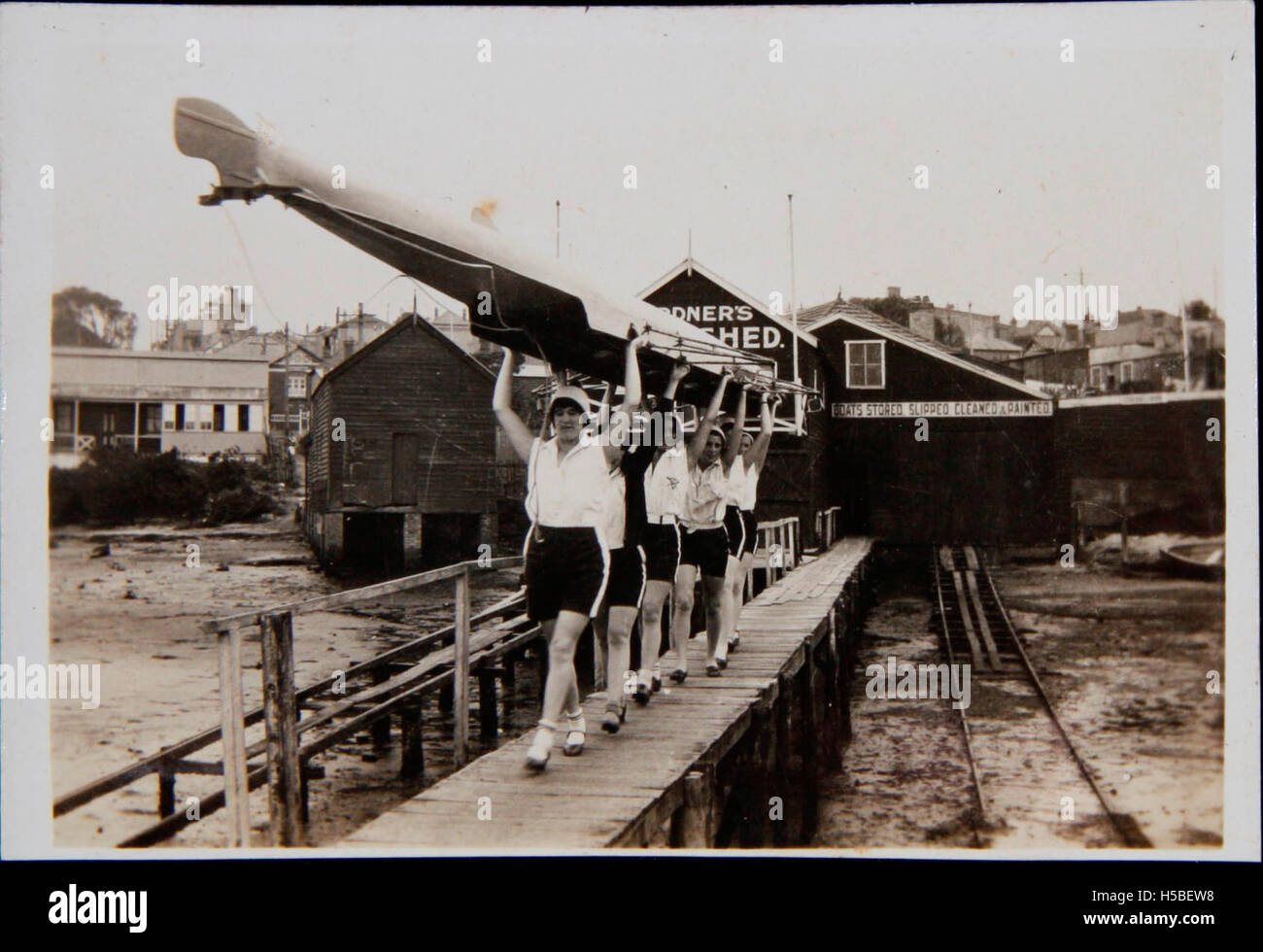 I membri della squadra di canottaggio femminile della YWCA vengono visti trasportare la loro barca dal capannone Gardner's Boat Shed, probabilmente una scena di un evento di canottaggio o di una sessione di allenamento. Questa immagine mette in evidenza le donne nello sport e la loro partecipazione al canottaggio durante i primi anni del XX secolo. Foto Stock