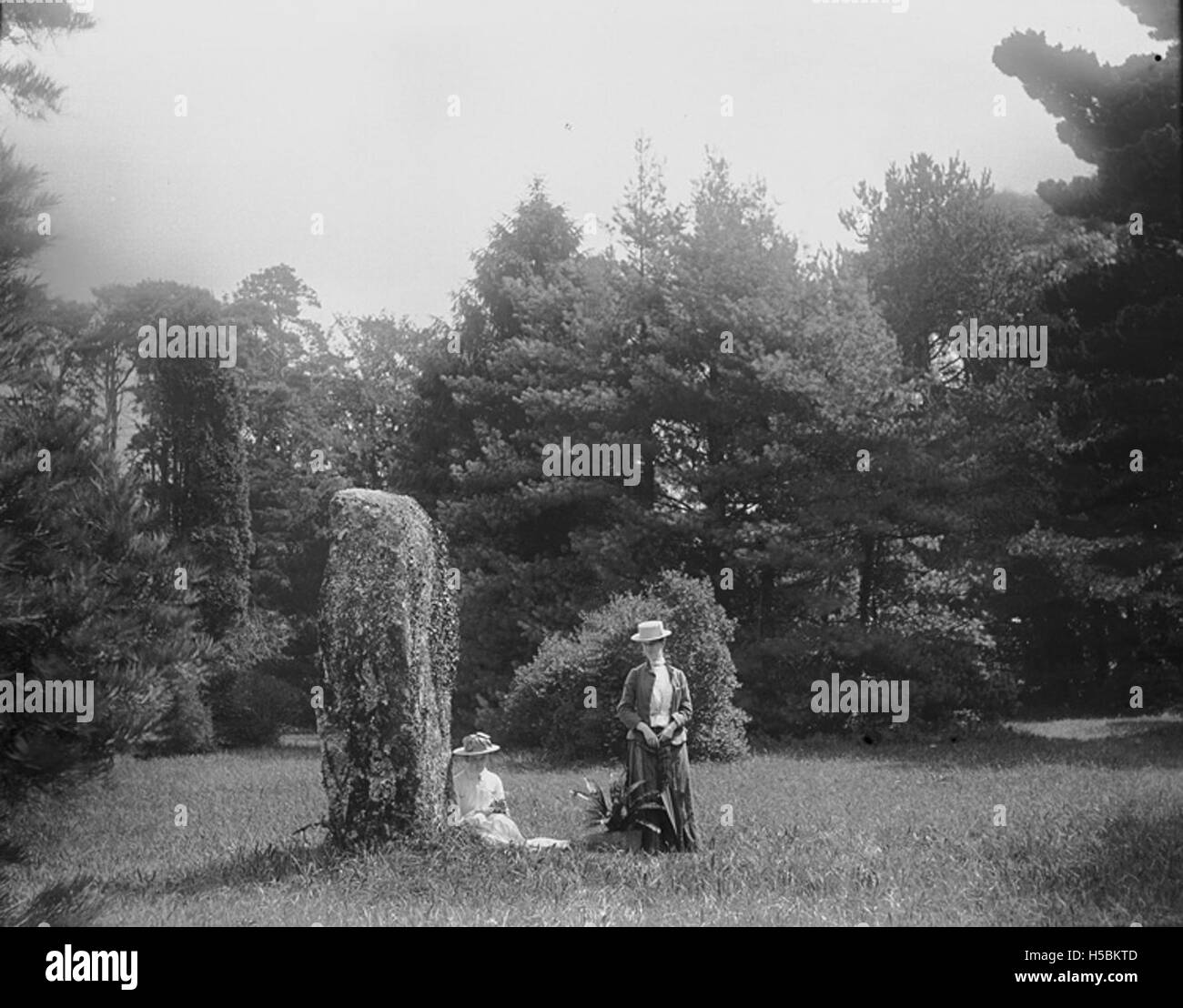 Questa immagine presenta due donne in piedi vicino a una pietra in piedi, forse un monumento preistorico o antico. Le pietre erette sono spesso associate a rituali storici o a significati culturali, in particolare nelle culture celtiche e preistoriche. Foto Stock