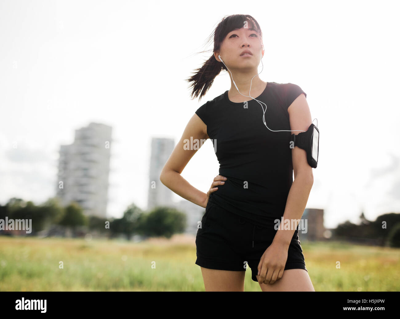 Colpo all'aperto del femminile in piedi nel parco urbano. Donna cinese in sportswear con gli auricolari per ascoltare musica e guardare Foto Stock