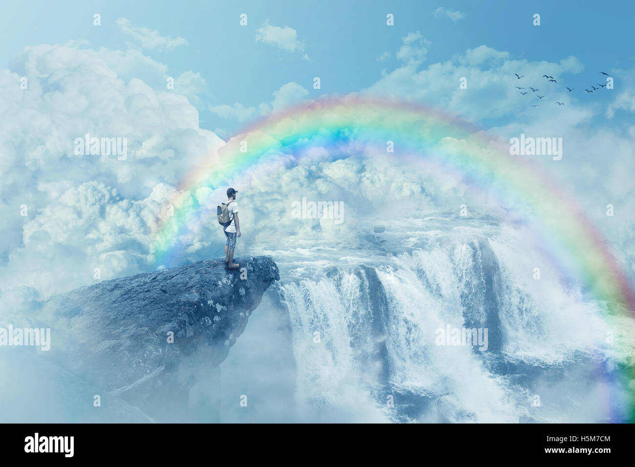 Giovane ragazzo su una scogliera guardando l'orizzonte con una cascata sopra le nuvole. Cammino di vita al di sotto di un arcobaleno in paradiso. Ispirazione Foto Stock