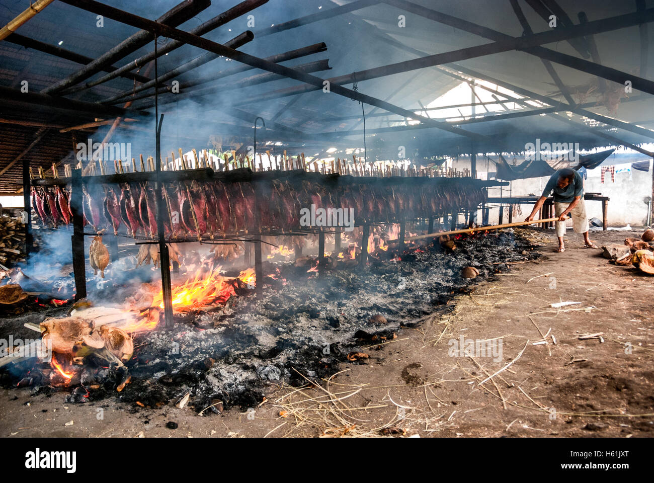 Un lavoratore sta mantenendo la distribuzione di buccia di cocco che brucia che è usato come combustibile per fumare le carni di tonno di skipjack in un'industria domestica in Indonesia. Foto Stock