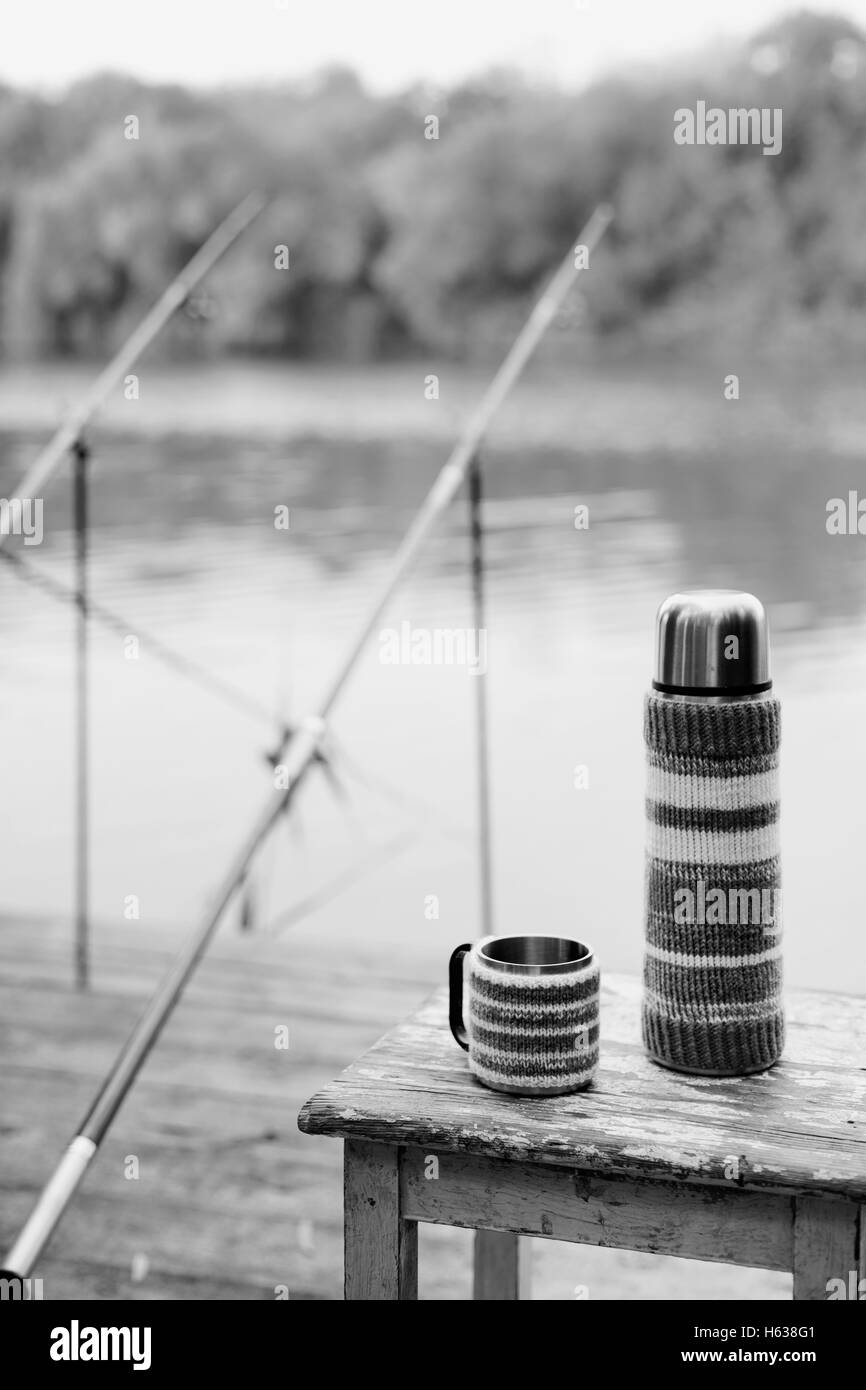 In bianco e nero photo. Pesca sul fiume. Una canna da pesca e un thermos con coppe in maglia copre. Foto Stock