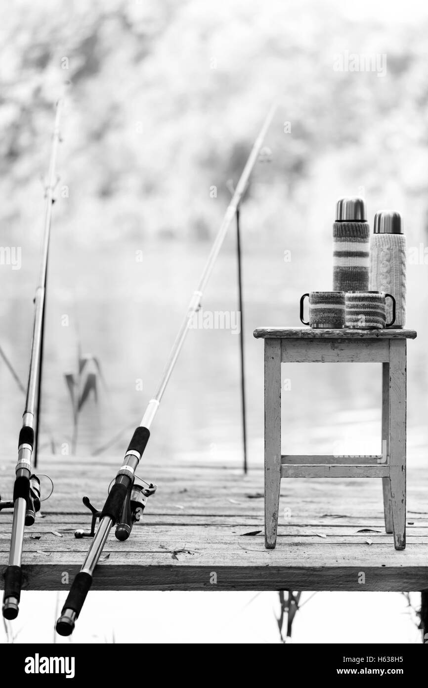 In bianco e nero photo. Pesca sul fiume. Una canna da pesca e un thermos con coppe in maglia copre. Foto Stock