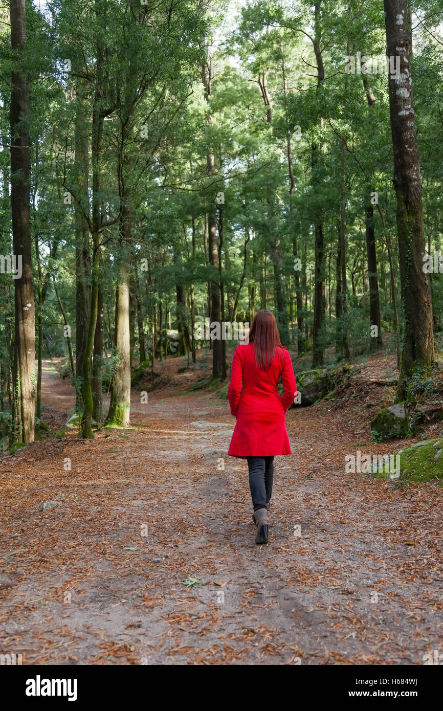 Giovane donna camminare da solo su un sentiero di bosco con un rosso lungo cappotto /// boschi lonely soprabito camicia legno a piedi passeggiando vagare lontano andando indietro Foto Stock