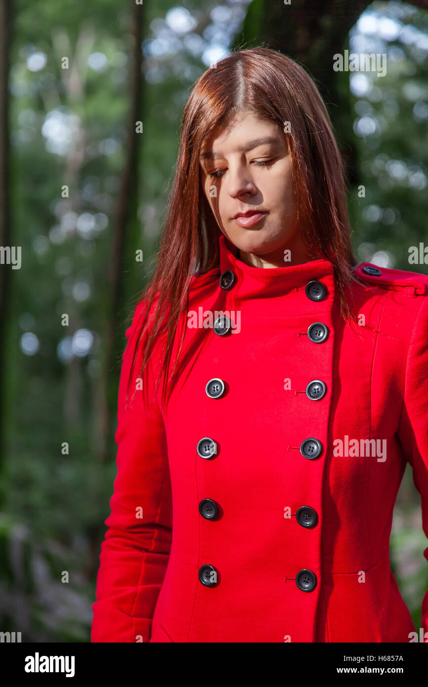 Giovane donna triste sensazione di camminare su una foresta che indossa un cappotto rosso in inverno sotto un raggio di luce solare Foto Stock