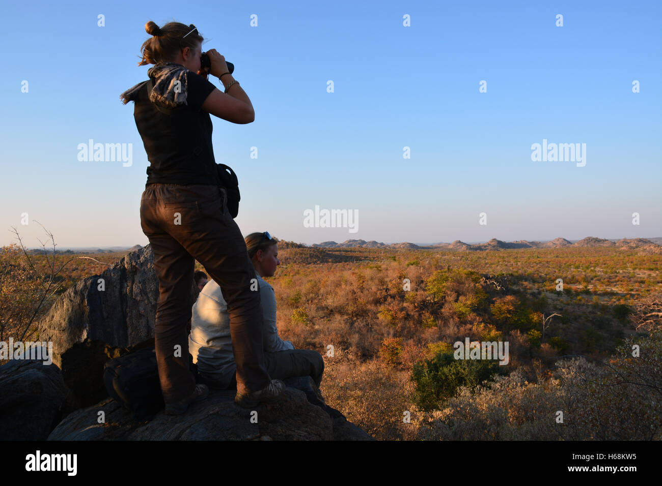 Una coppia di giovani studenti volontari donna sondaggio il paesaggio al tramonto dalla cima di un kopje nel deserto boccola del Botswana, Africa. Foto Stock