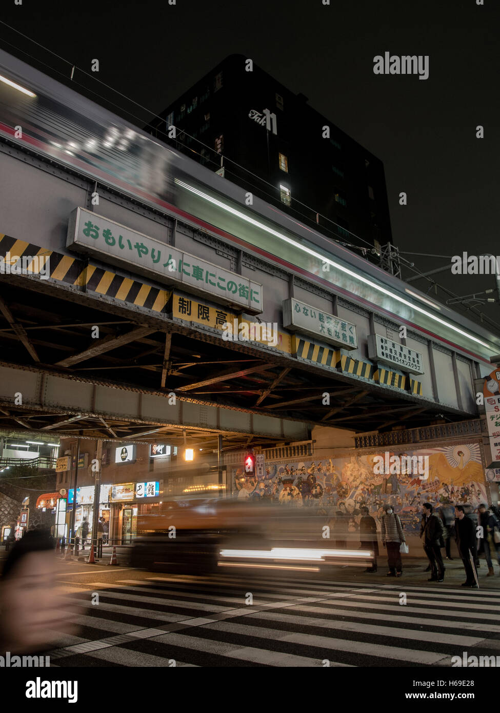 Tokyo di notte. Vetture blur passato pedoni in attesa in corrispondenza del punto di attraversamento al di fuori della stazione Takadanobaa come una linea Yamanote treno passa overhead. Foto Stock