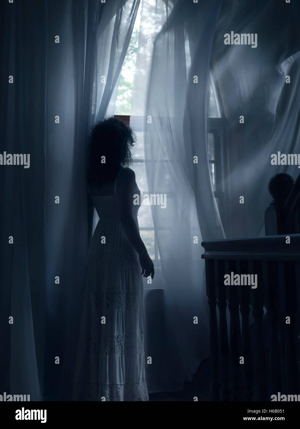 Licenza e stampe a MaximImages.com - giovane donna in un vestito bianco in piedi da una finestra con volare nelle tende del vento in una stanza buia di una casa Foto Stock
