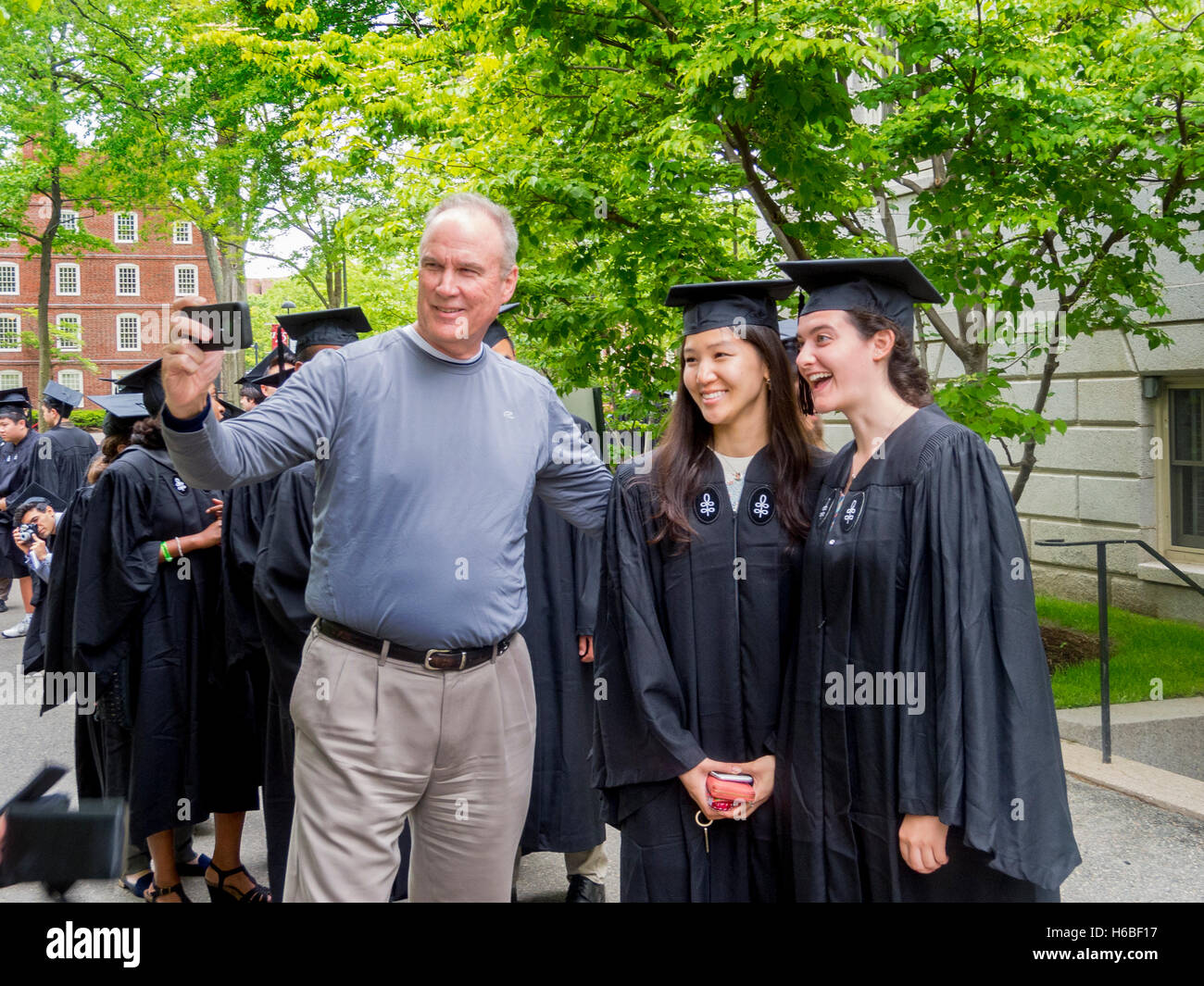 Usando un telefono cellulare fotocamera, un orgoglioso padre prende un selfie foto di se stesso con la sua figlia e suo Asian migliore amico in tappi di graduazione e gli abiti sul giorno di graduazione presso la Harvard University di Cambridge, MA. Foto Stock