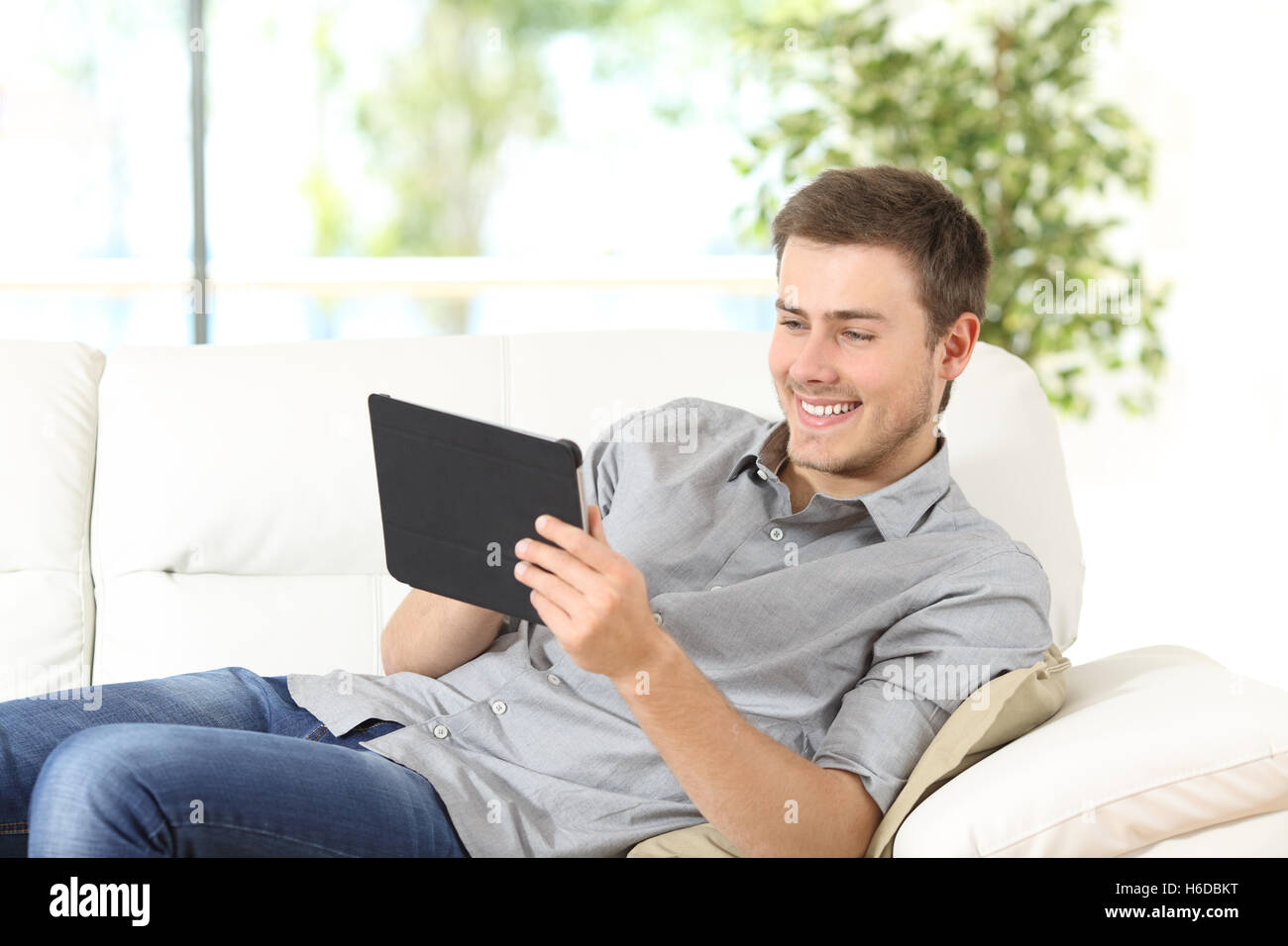Felice l'uomo utilizzando una compressa sulla linea seduto su un divano in soggiorno a casa con una finestra in background Foto Stock