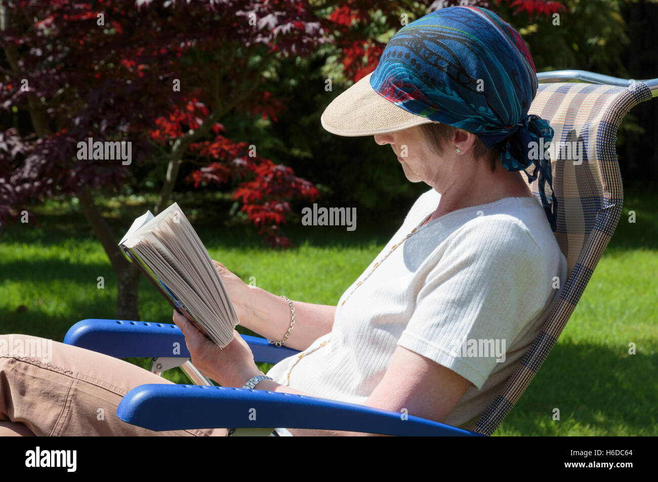 Qualcuno si è ritirato senior donna rilassante in un cortile sedia da giardino leggendo un libro in estate sole ombreggiata da cappello. Stile di vita Hygge sulla giornata di sole. Regno Unito Foto Stock