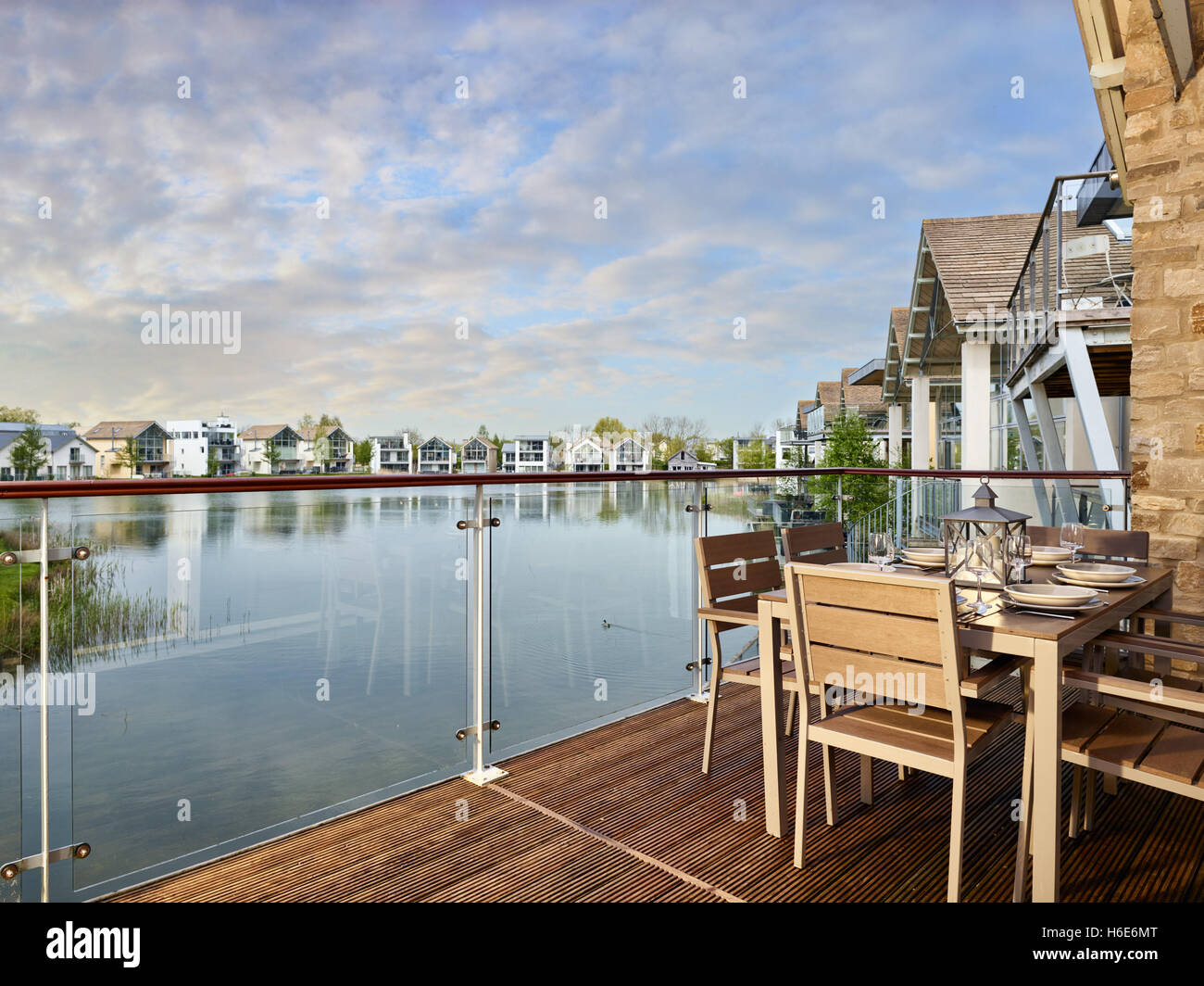 Un set tavolo da pranzo e sedie sul decking affacciato su un lago circondato da case vacanza su una bella serata in Cotswolds, Regno Unito Foto Stock