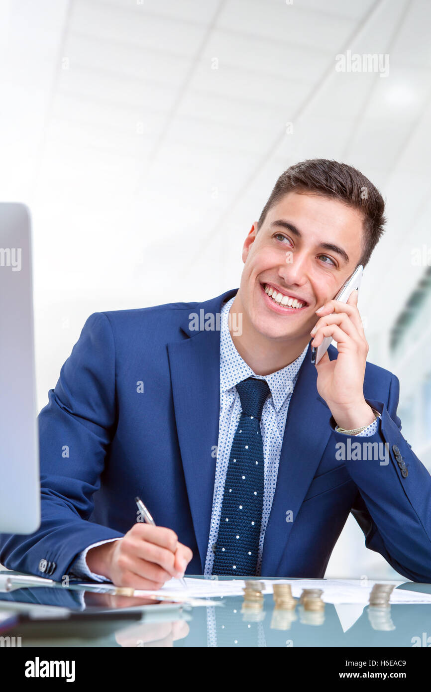 Close up ritratto del bel giovane imprenditore parlando su smart phone in ufficio. Giovane uomo alla scrivania nella parte anteriore del computer. Foto Stock