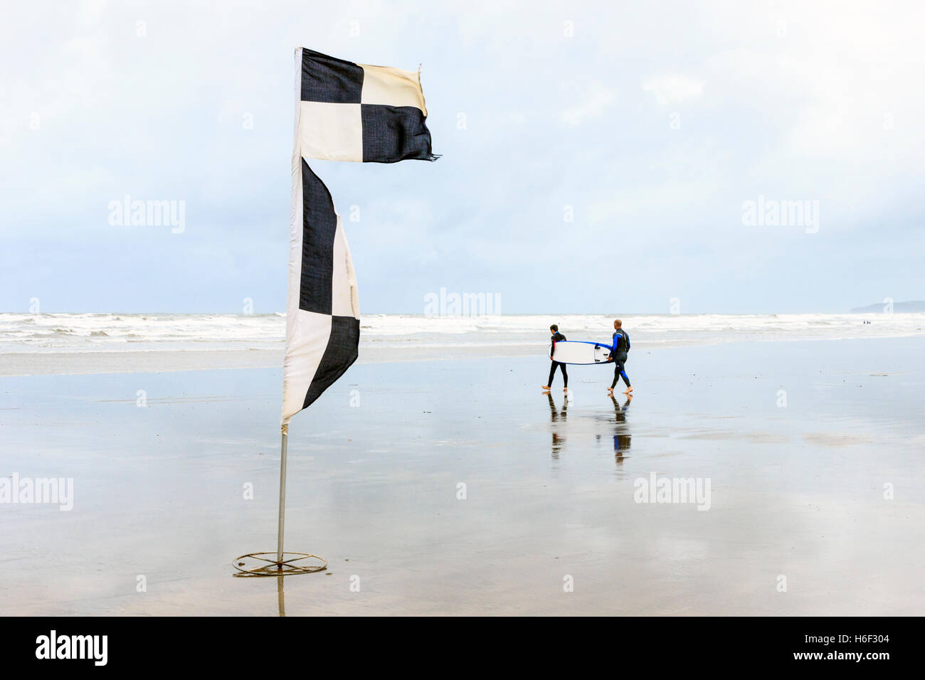 Due surfisti che porta una tavola da surf a mare con la bassa marea sulla spiaggia di Condino, Devon, Inghilterra, Regno Unito, un bianco e nero flag di controllo in primo piano Foto Stock