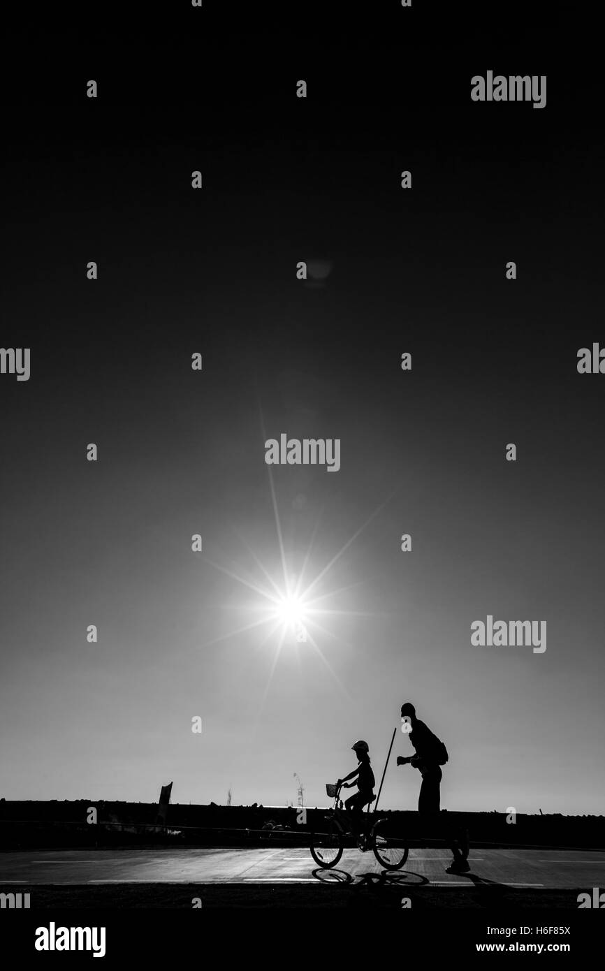 Silhouette di padre esegue da sua figlia in bicicletta, passando su un percorso del parco in una limpida giornata estiva, pomeriggio di sole splendente. Foto Stock