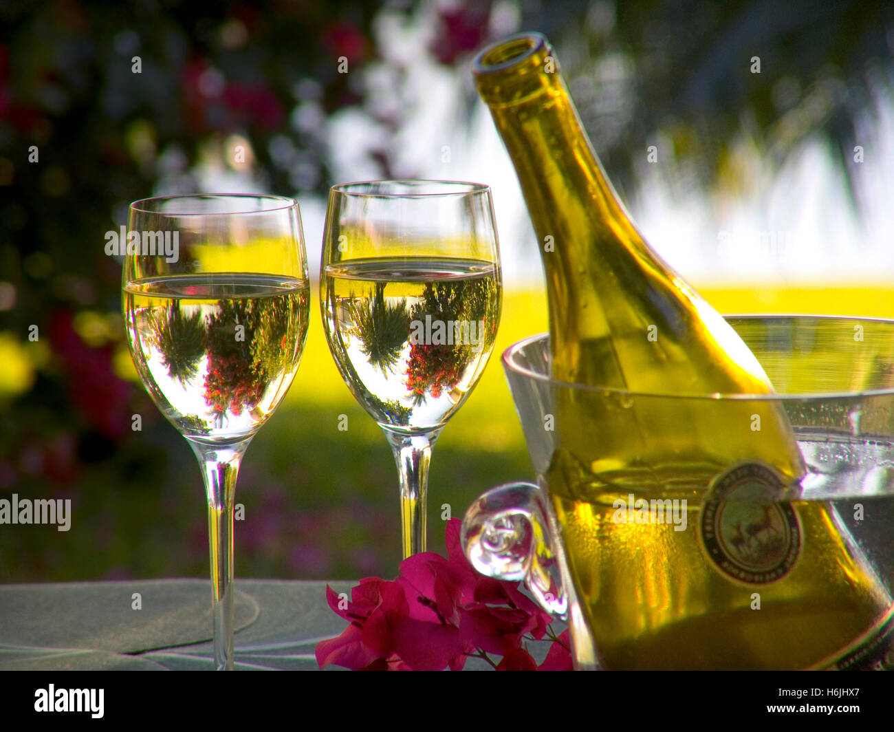 Bicchieri da vino bianco per le vacanze, bicchiere e bottiglia di cristallo tavolo da giardino all'aperto all'aperto con fiori di bougainvillea e palma alberi dietro in un ambiente di vacanza esotico di lusso Foto Stock