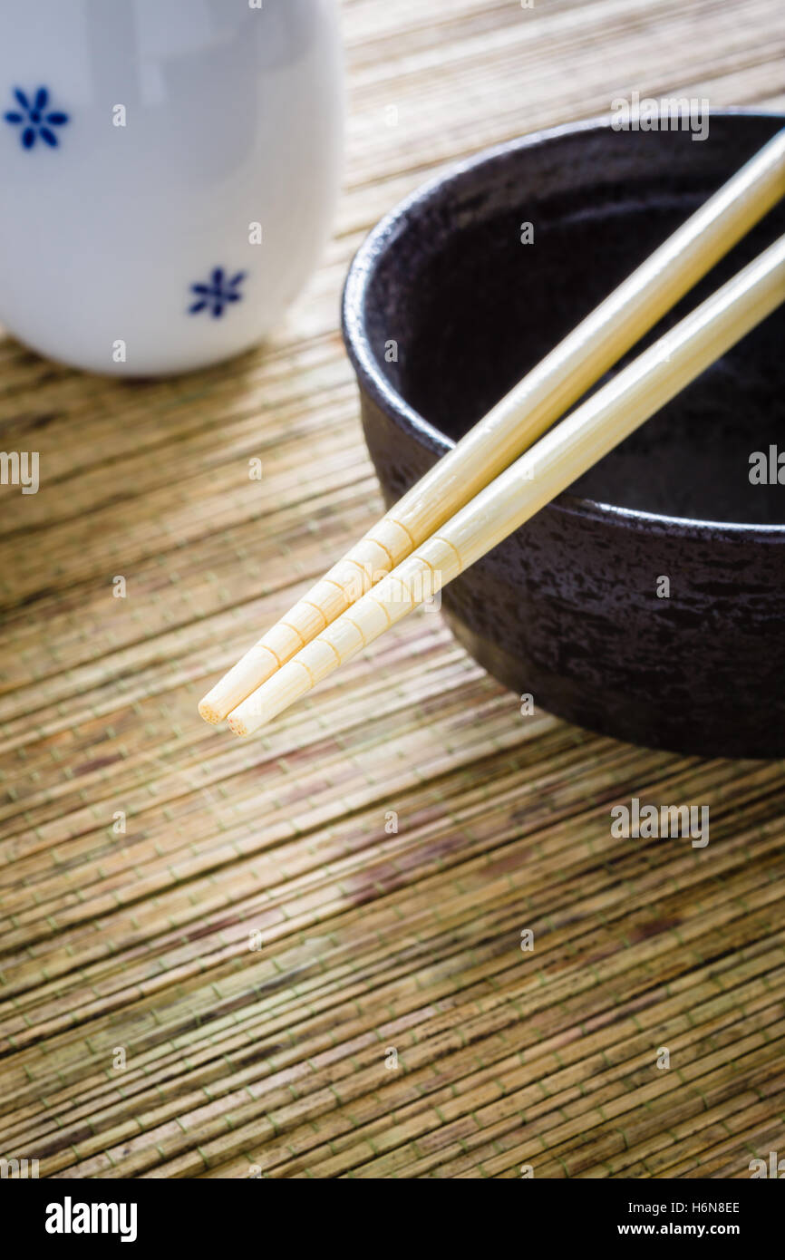 Bacchette e Riso o Noodle ciotola con una bottiglia di sake contro un bambù tappetino di messa a fuoco lo sfondo su bacchette Foto Stock
