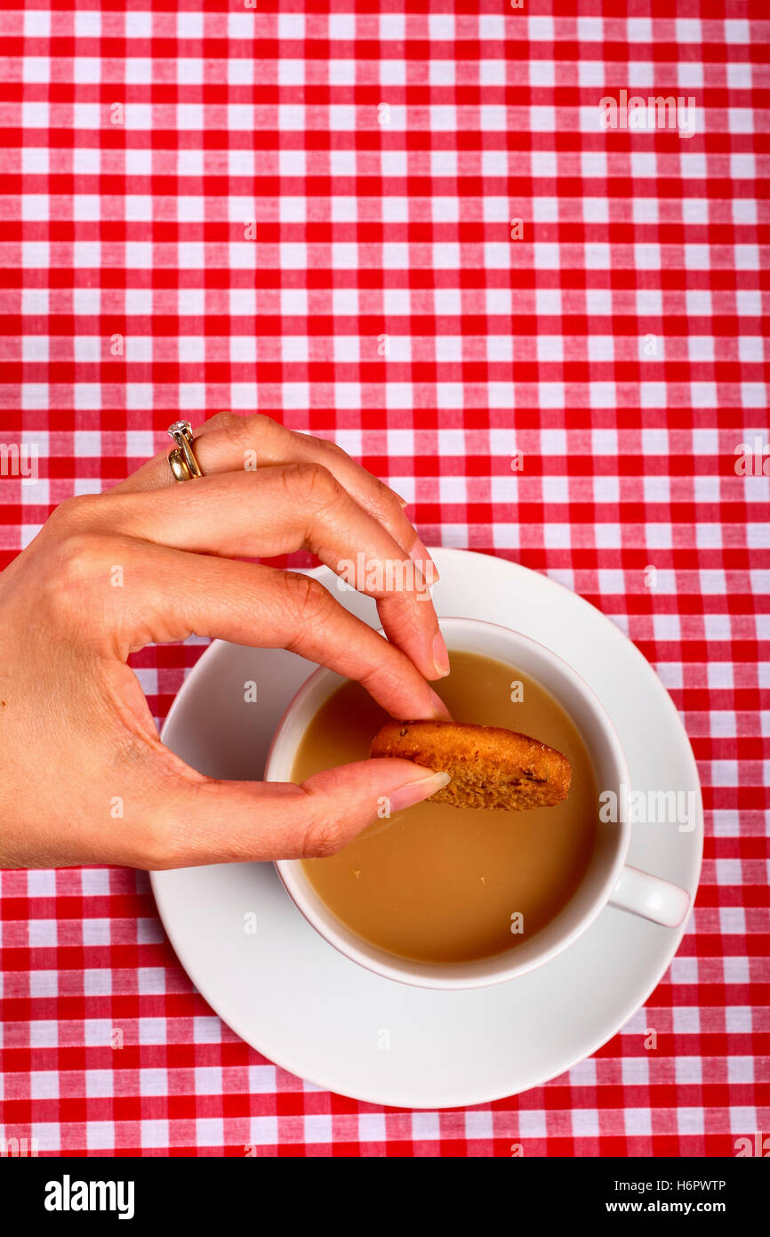 Donna-ladies-ragazze dunking mano un cookie biscotto in una calda tazza di tè con un punto rosso e bianco a scacchi tabella coperta di stoffa Foto Stock