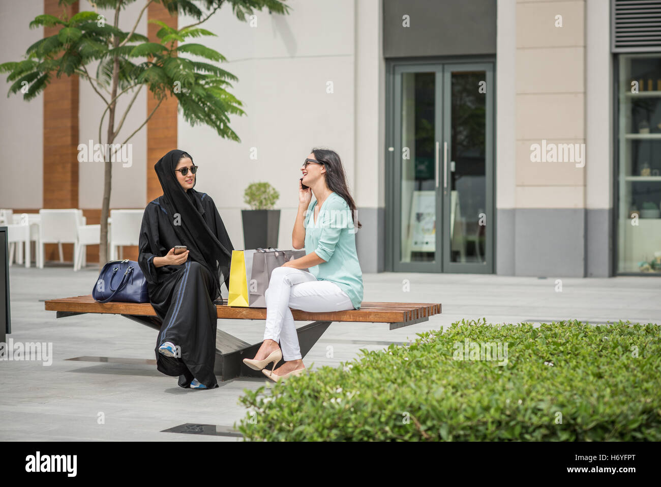 Giovani medio orientale donna che indossa abbigliamento tradizionale seduta sul banco di lavoro con amico femmina, Dubai, Emirati Arabi Uniti Foto Stock