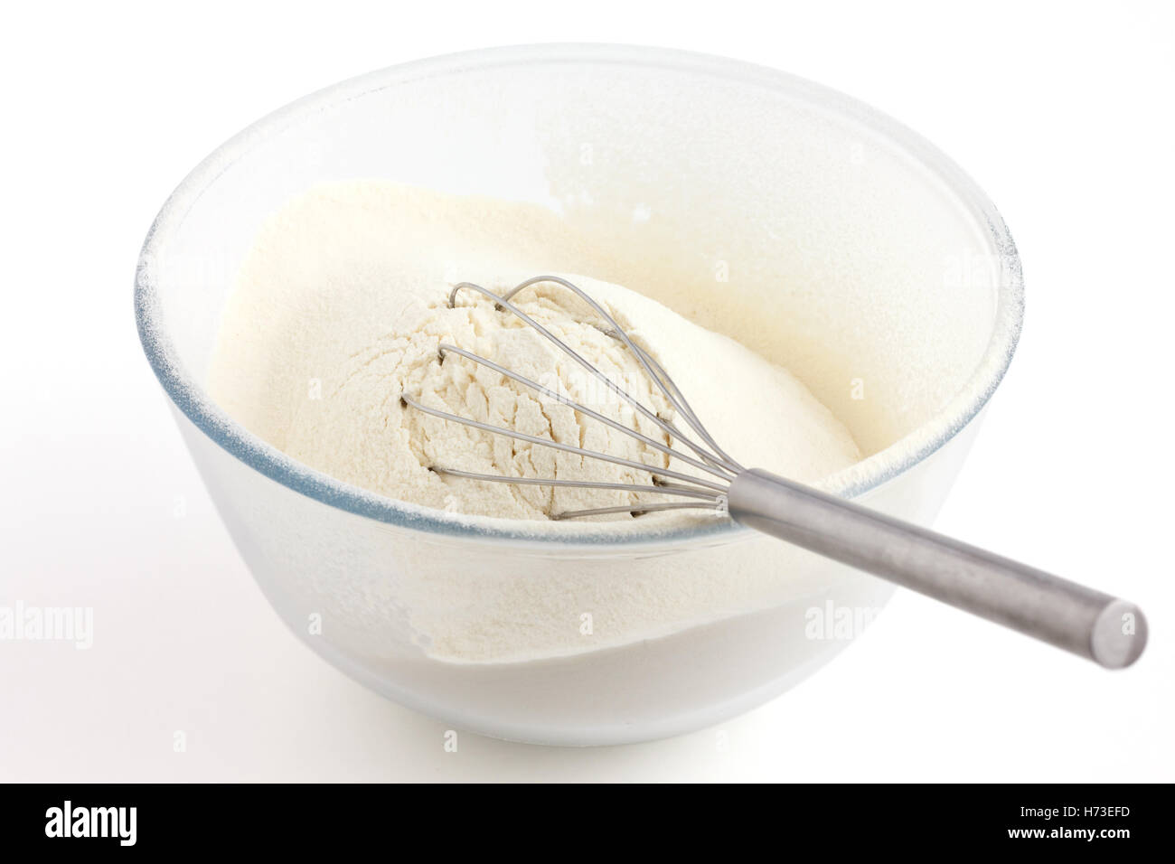 Mescolare con una frusta giacente in farina di frumento bianca in un piatto di vetro. Sfondo bianco. Foto Stock