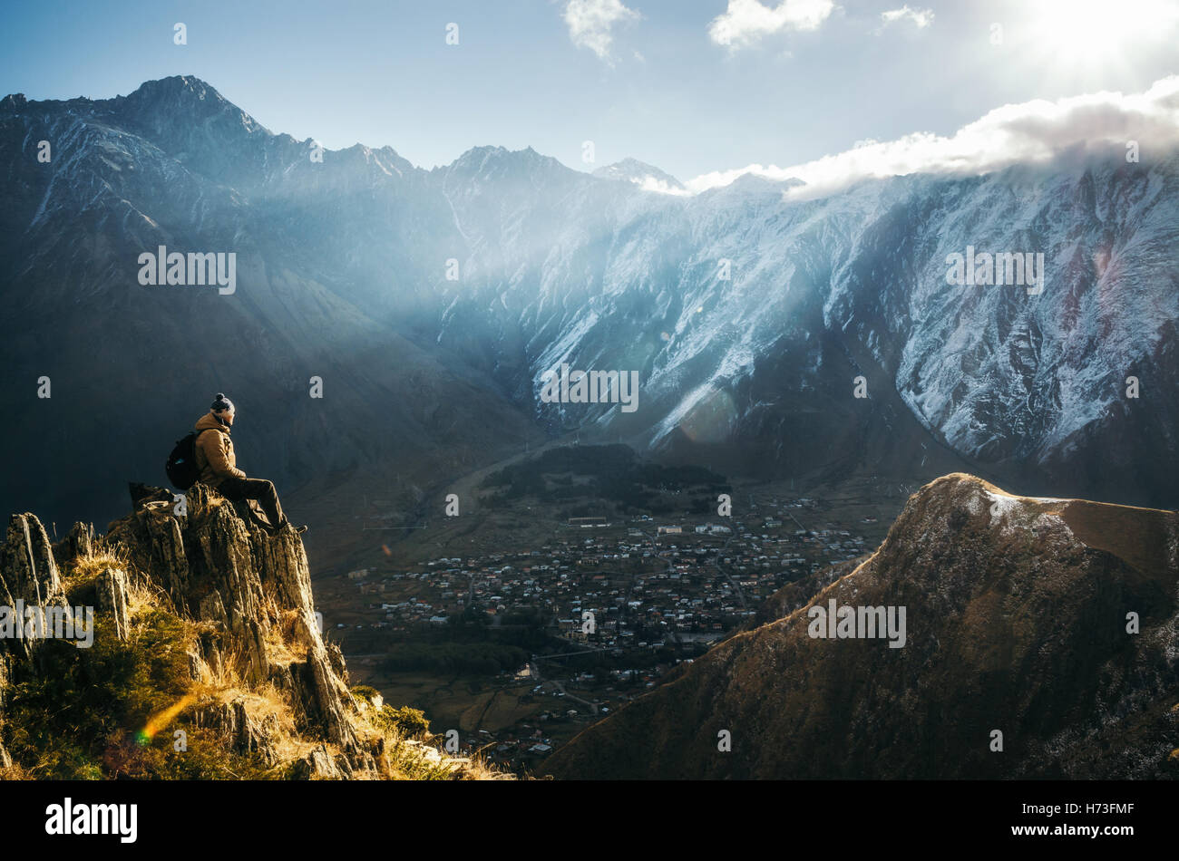 Giovani turisti in bright hat, pantaloni neri con uno zaino sedersi sulla scogliera di bordo e guardando il misty mountain village Foto Stock