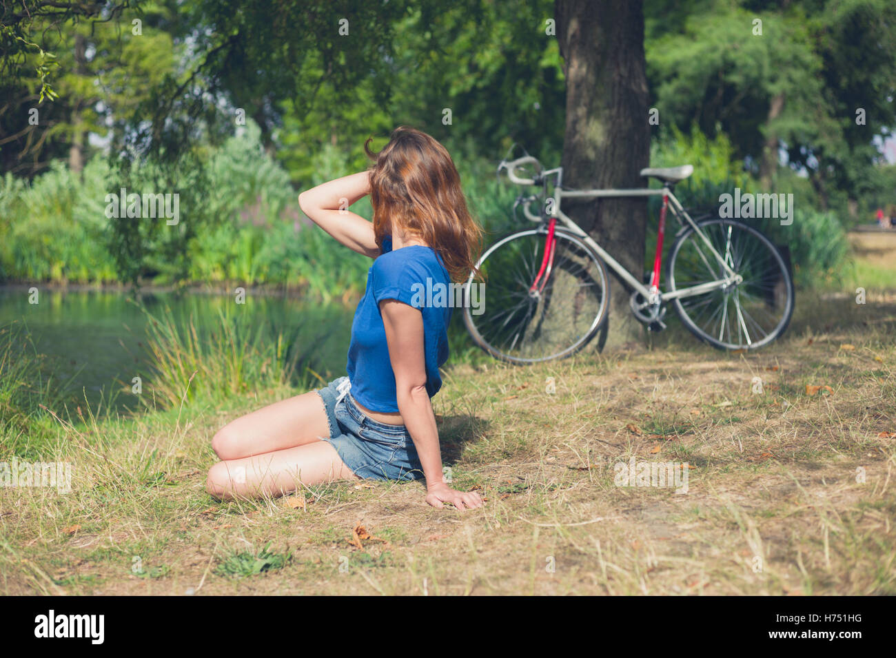Una giovane donna è rilassante dall'acqua in un parco in un giorno d'estate con la sua bicicletta in appoggio contro un albero in background Foto Stock
