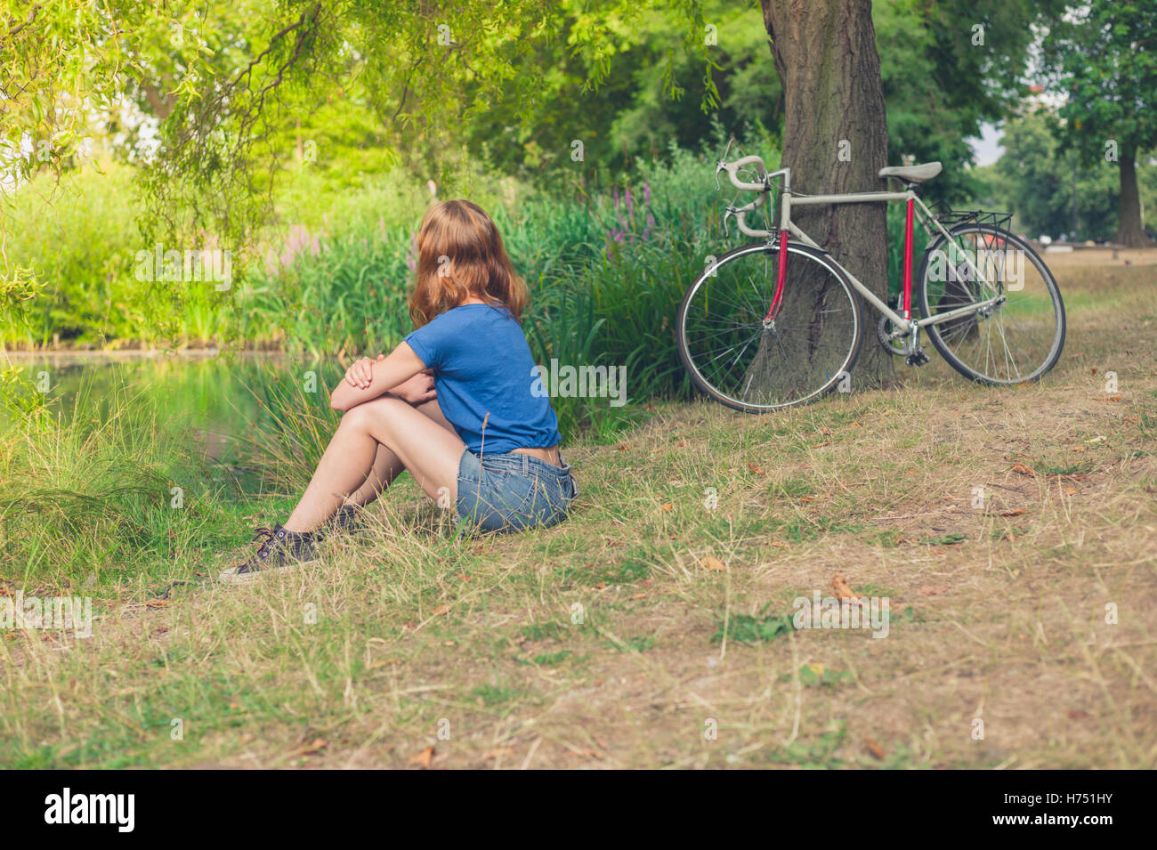 Una giovane donna è rilassante dall'acqua in un parco in un giorno d'estate con la sua bicicletta in appoggio contro un albero in background Foto Stock