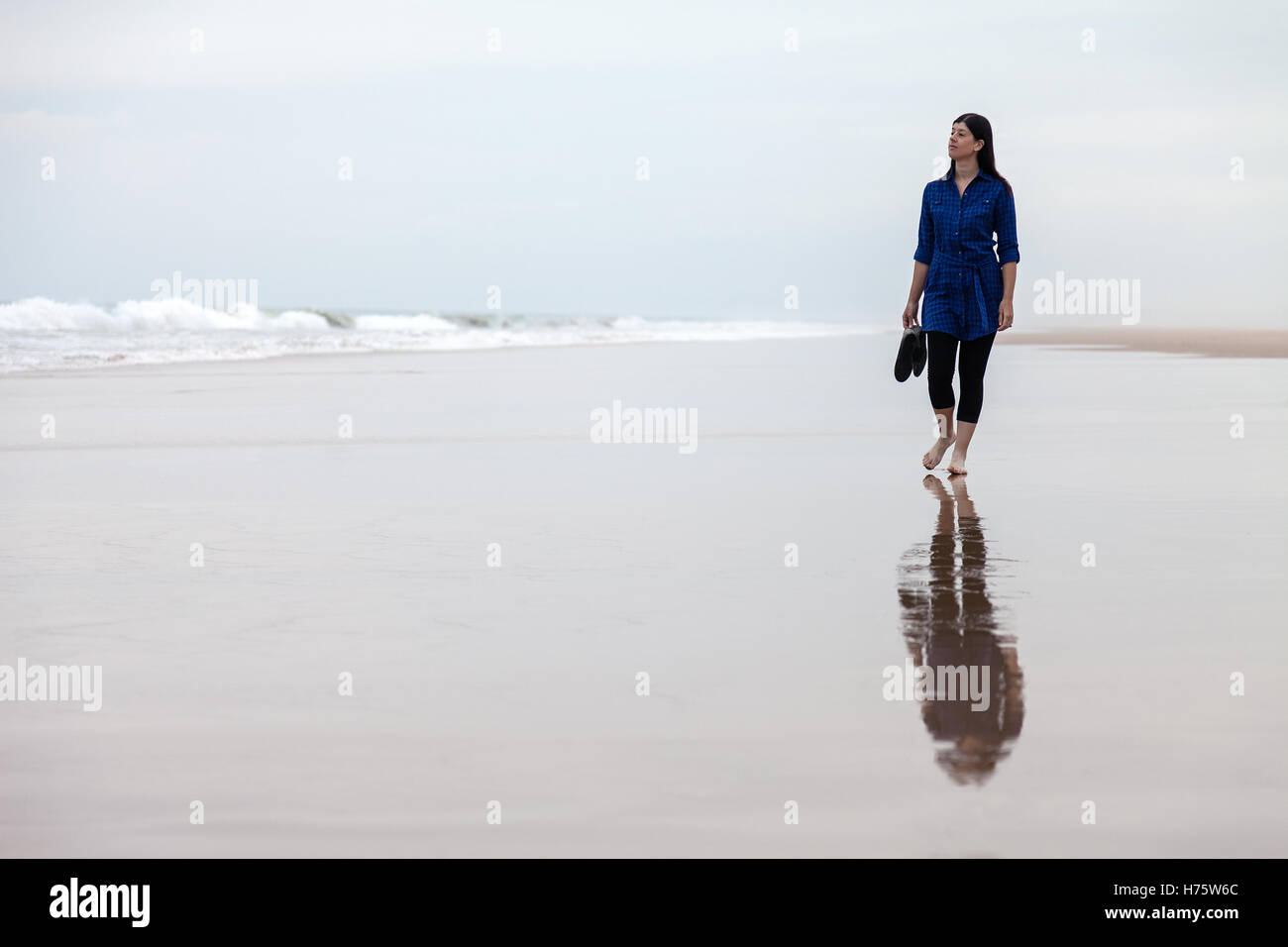 Giovane donna camminare da solo in una spiaggia deserta riflessa sulla sabbia bagnata in una giornata autunnale. Foto Stock