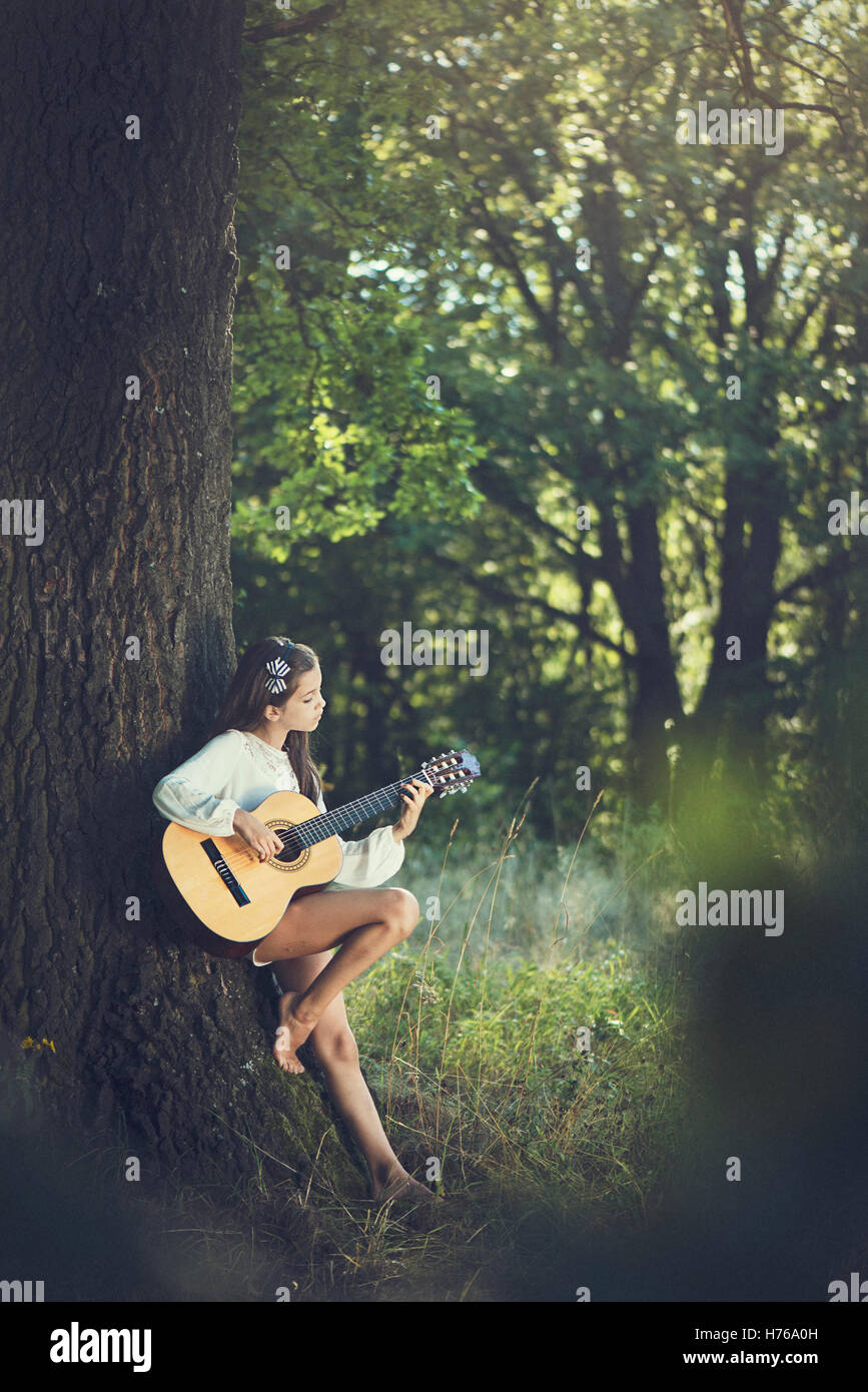 Ragazza appoggiata contro un albero a suonare la chitarra Foto Stock
