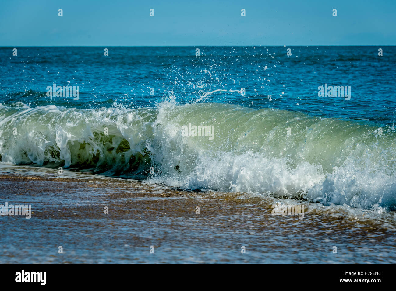 Rottura d'onda su un litorale di sabbia con battenti spray e una calma oceano blu sullo sfondo Foto Stock