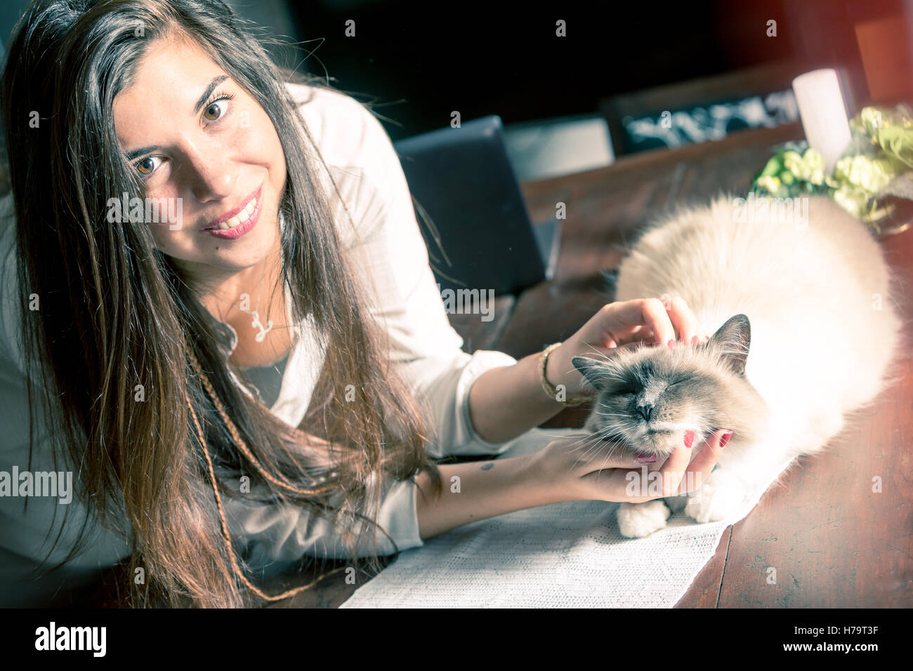Attraente donna felice rilassante con il suo gatto di casa che accarezzano la sua testa come si rilassa al sole su di un tavolo da pranzo, chiudere vie Foto Stock