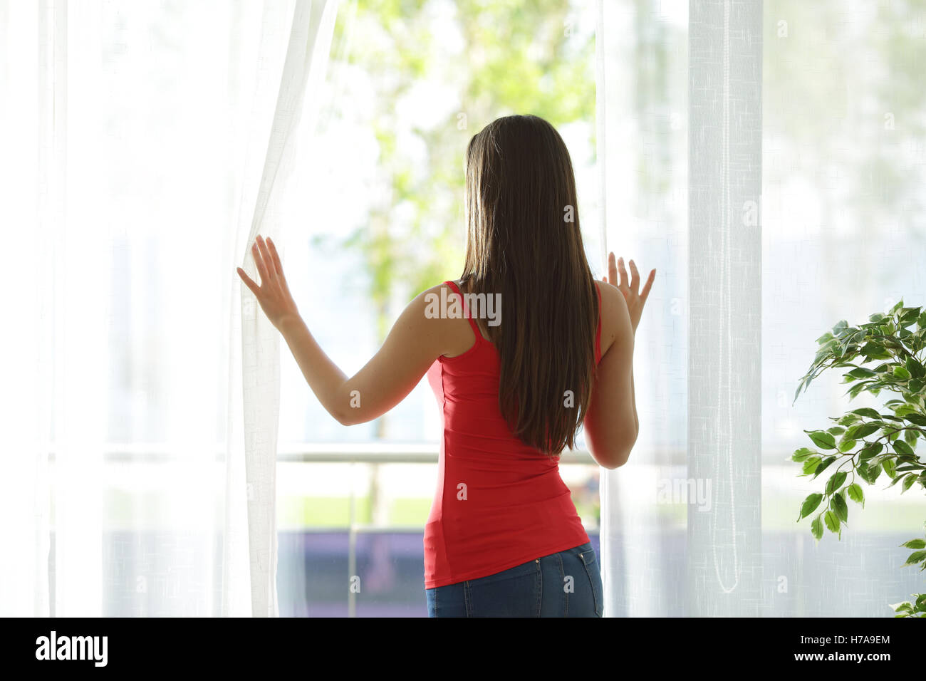 Vista posteriore di una donna che guarda all'esterno attraverso una finestra e aprire le tende a casa Foto Stock