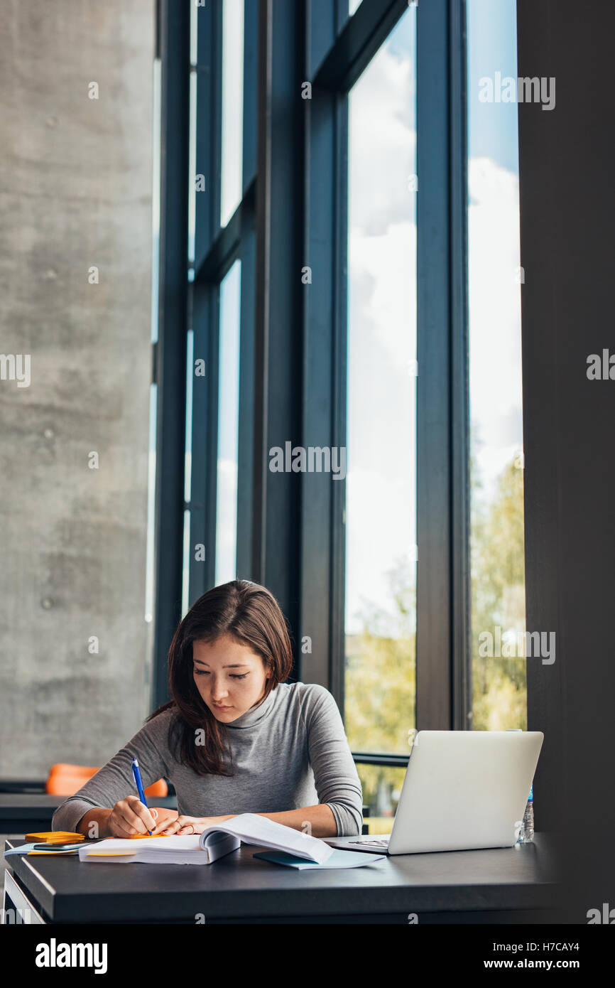 Studentessa prendendo appunti da libri per il suo studio. Giovane donna seduta a tavola con libri in biblioteca universitaria. Foto Stock
