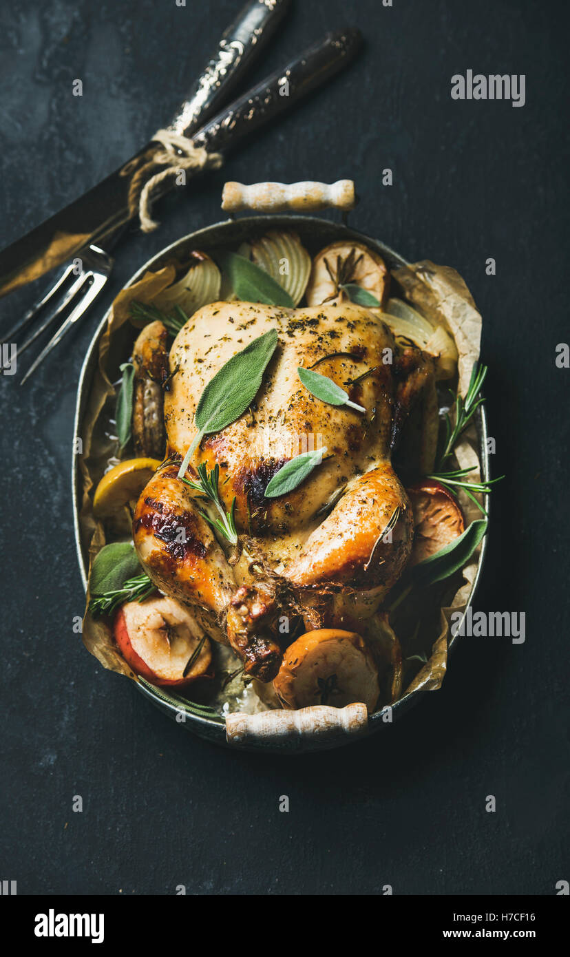 Arrosto di pollo intero con cipolla, mele e salvia in vassoio con posate su pietra scura sullo sfondo, vista dall'alto, selezionare Foto Stock