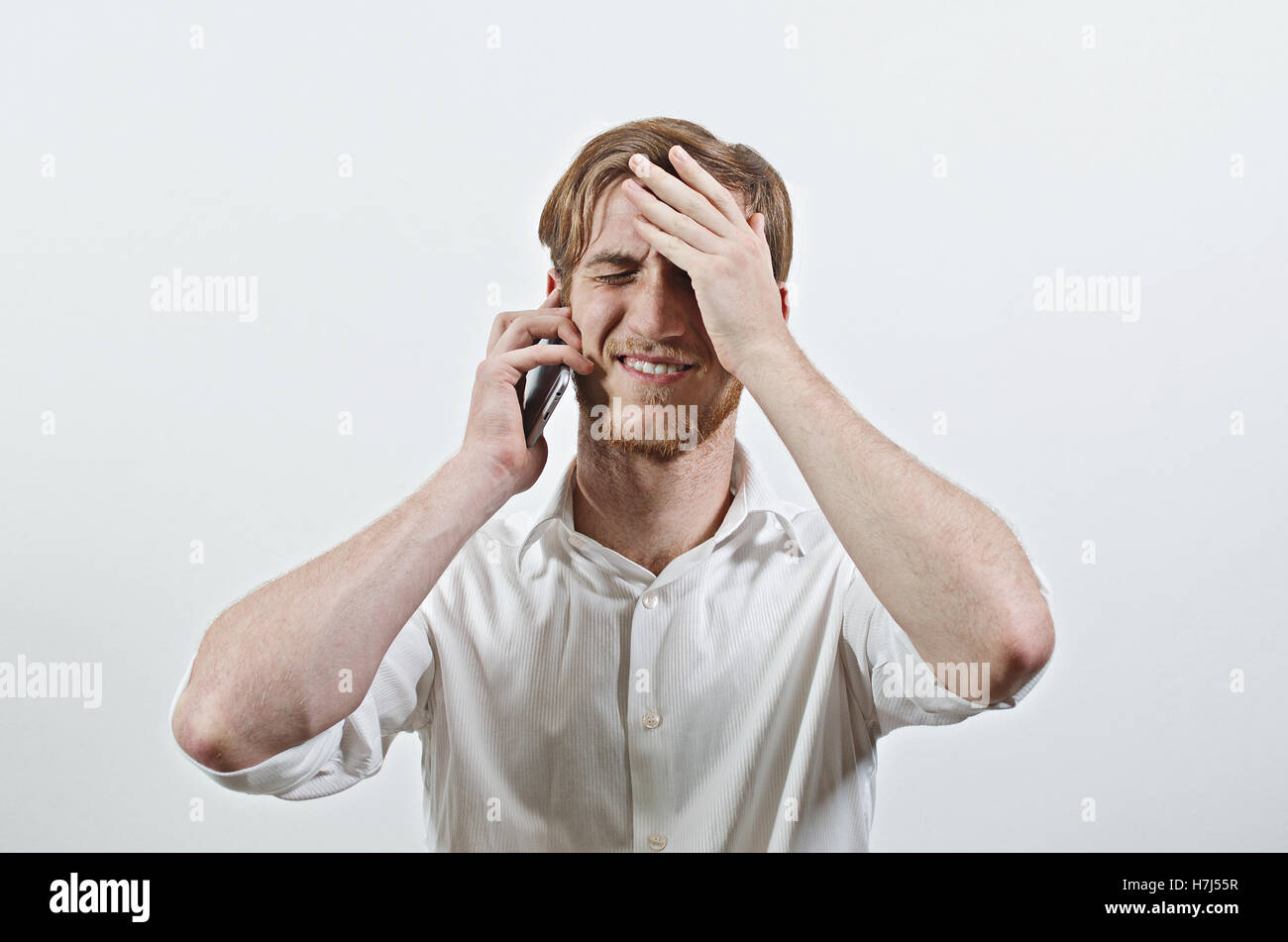 Giovane Adulto Uomo in camicia bianca ascoltando il suo telefono, tenendo la testa tra le mani, ricevere cattive notizie Foto Stock